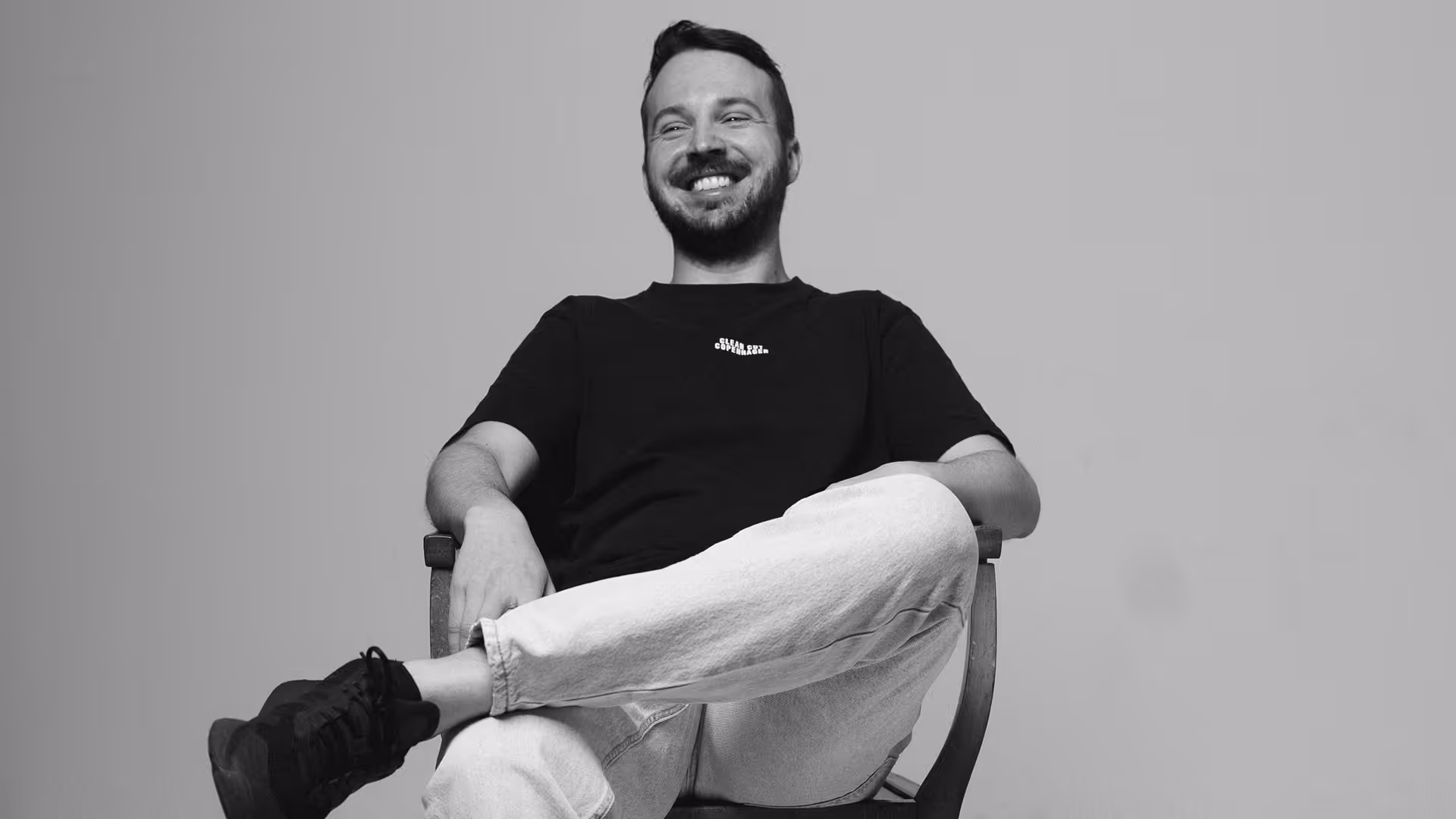 Ein lächelnder Mann sitzt entspannt auf einem Stuhl mit verschränkten Beinen vor einem hellen Hintergrund.