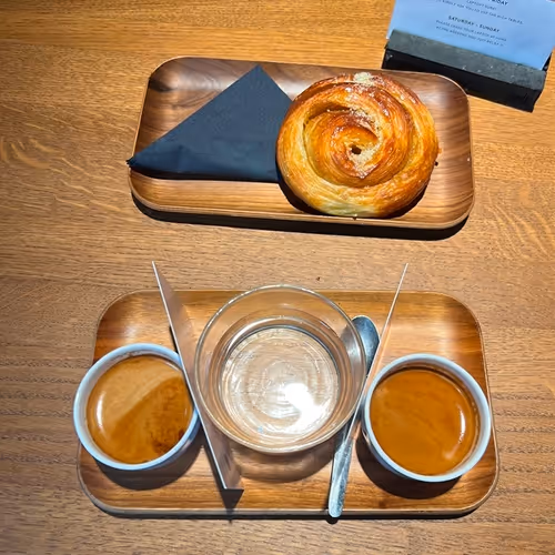 Zwei Espressi, ein Glas Wasser und ein Löffel auf einem Holztablett, daneben ein weiteres Tablett mit einem Gebäck und schwarzer Serviette auf Holztisch.