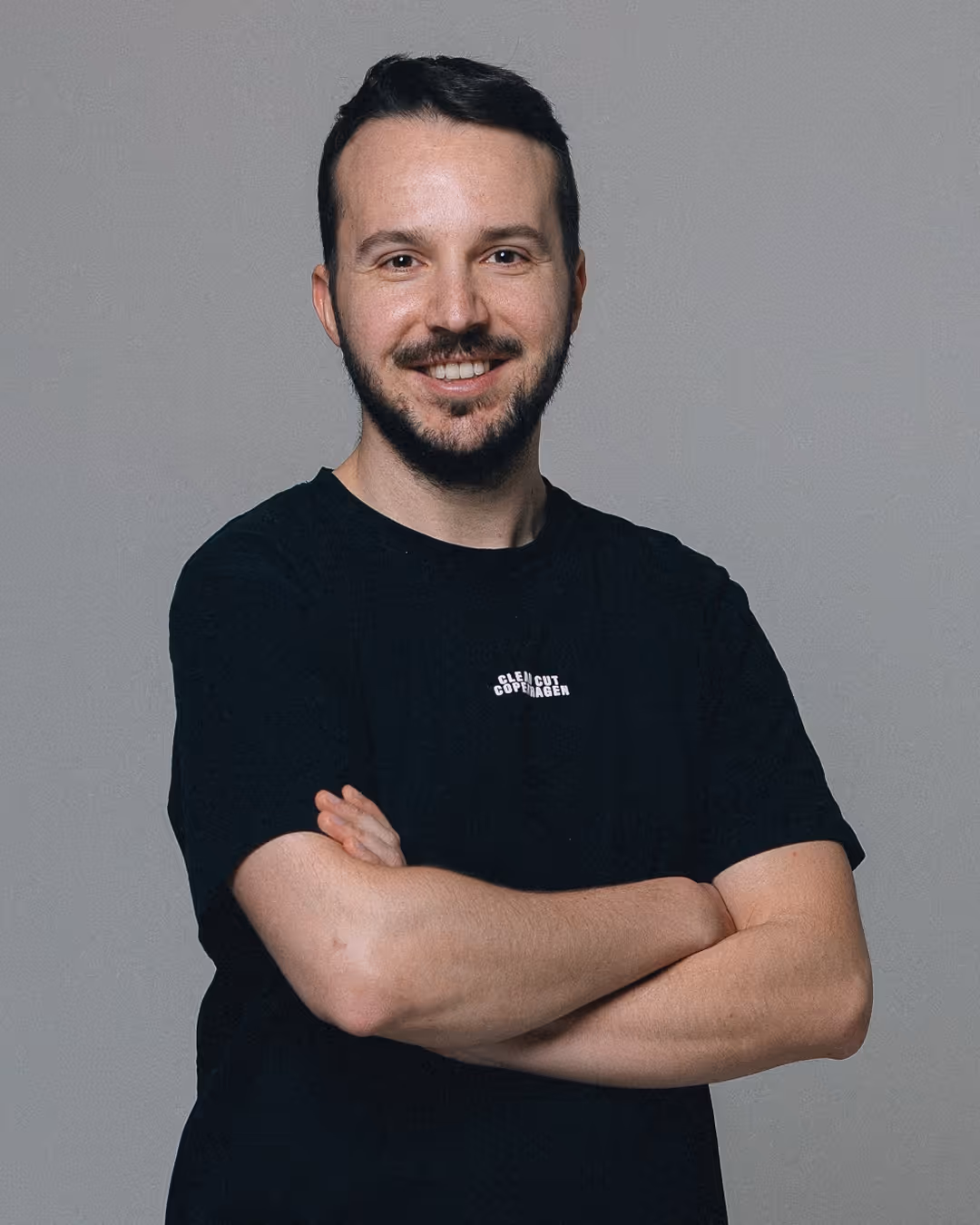 Junger Mann mit kurzen dunklen Haaren und Bart, der vor grauem Hintergrund lächelt und die Arme vor der Brust verschränkt trägt.