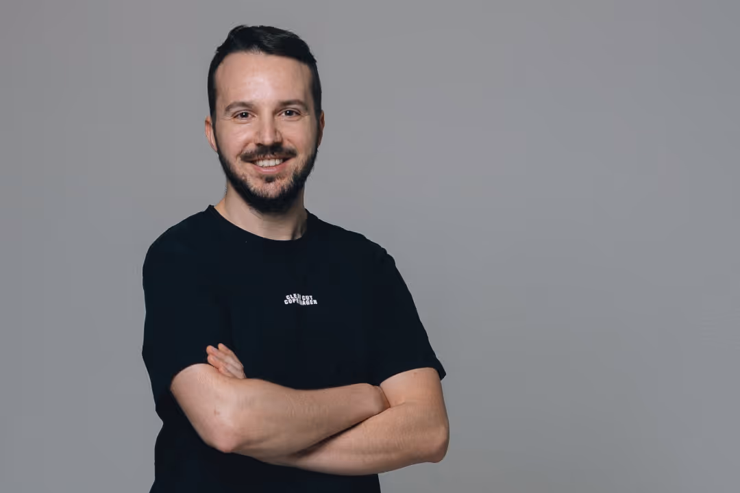 Ein lächelnder Mann mit dunklem Bart und kurzem Haar, der die Arme vor der Brust verschränkt, vor einem grauen Hintergrund.