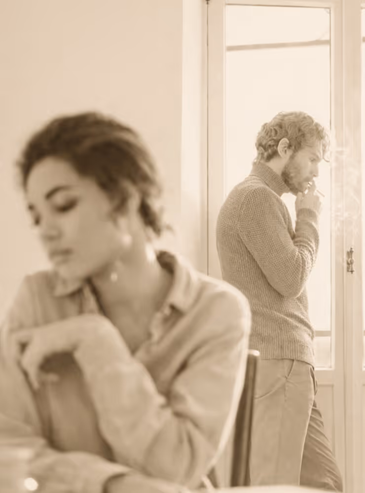 Een gepijnigde vrouw zit op de voorgrond met gesloten ogen die haar relatie wilt verbeteren terwijl haar man in de achtergrond bij het raam staat.