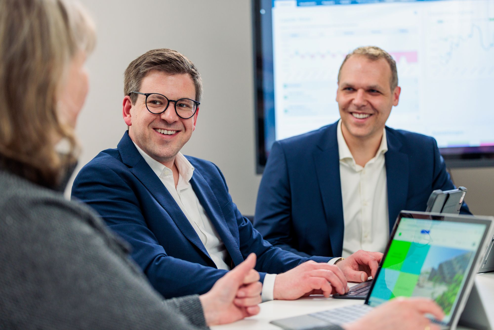 Zwei Männer in Businesskleidung sitzen an einem Besprechungstisch und lächeln.