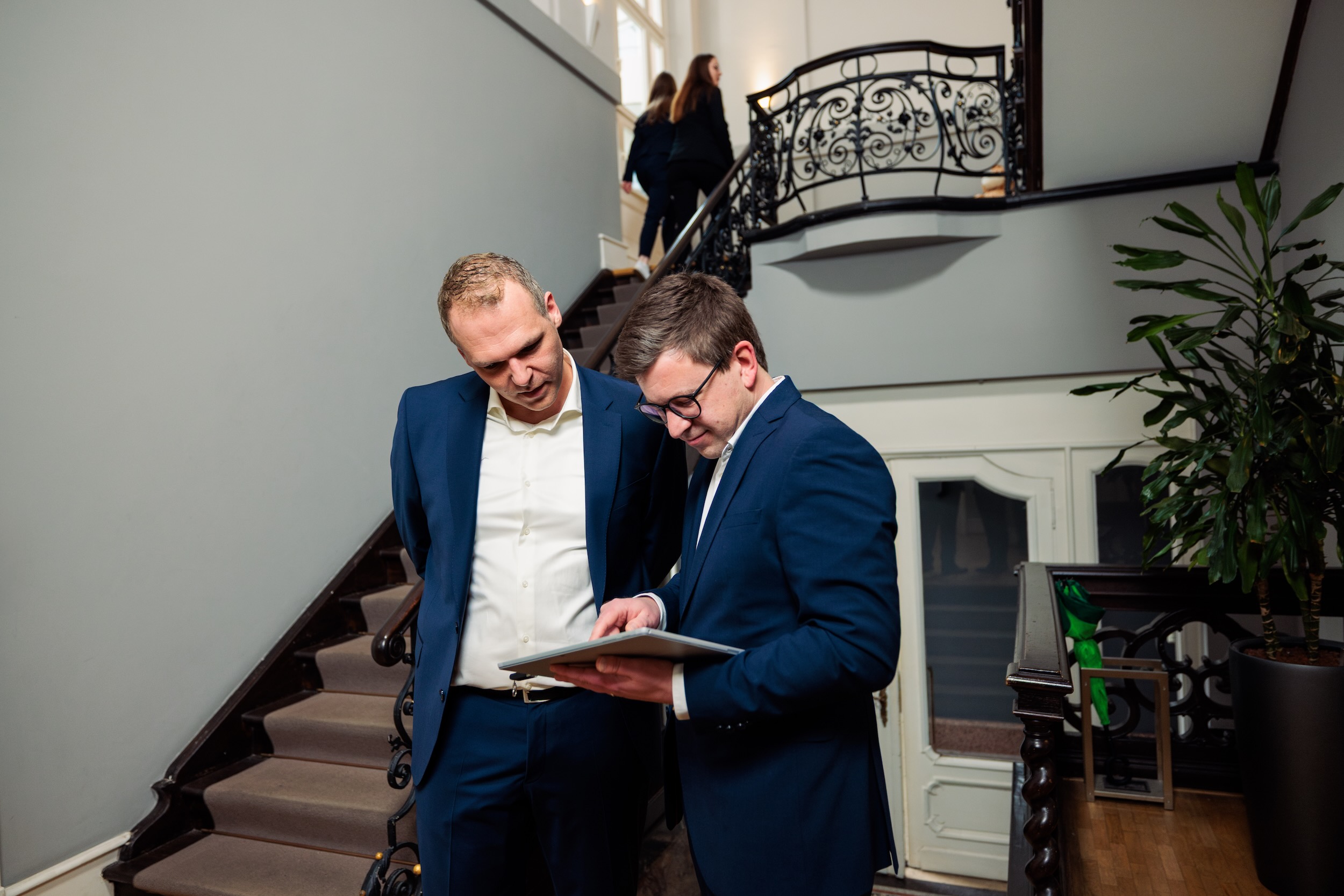 Zwei Männer stehen vor einer Treppe. Ein Mann zeigt dem anderen etwas auf einem Tablet.