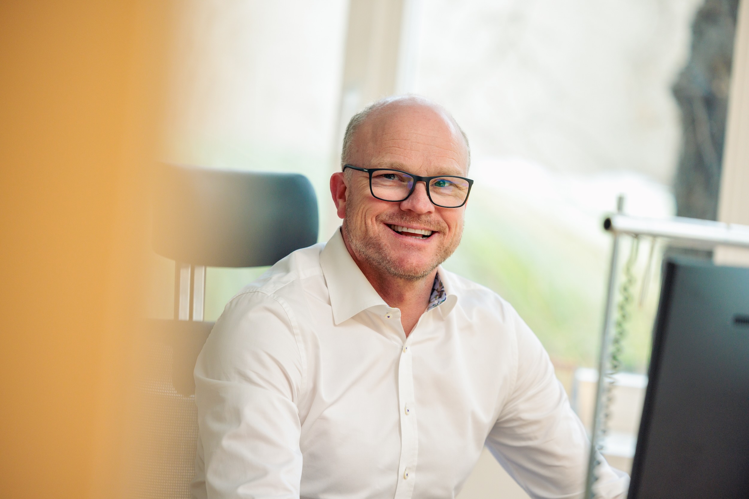 Ein Mann mit Brille und weißem Hemd sitzt an einem Schreibtisch und lächelt.