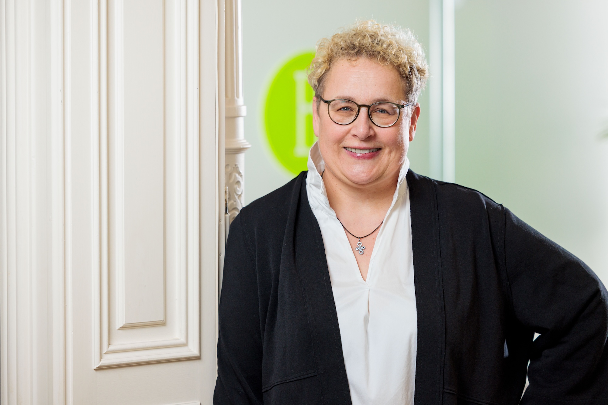 Porträt einer blonden Frau mit Brille und schwarzem Sakko.