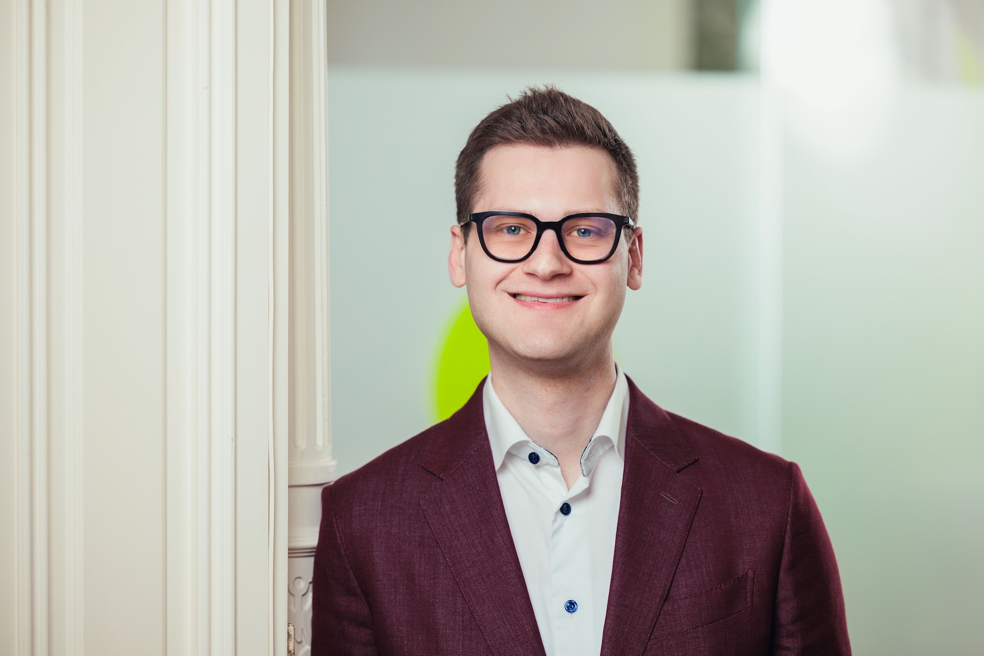 Porträt eines brünetten Mannes mit dunkelrotem Sakko und Brille.