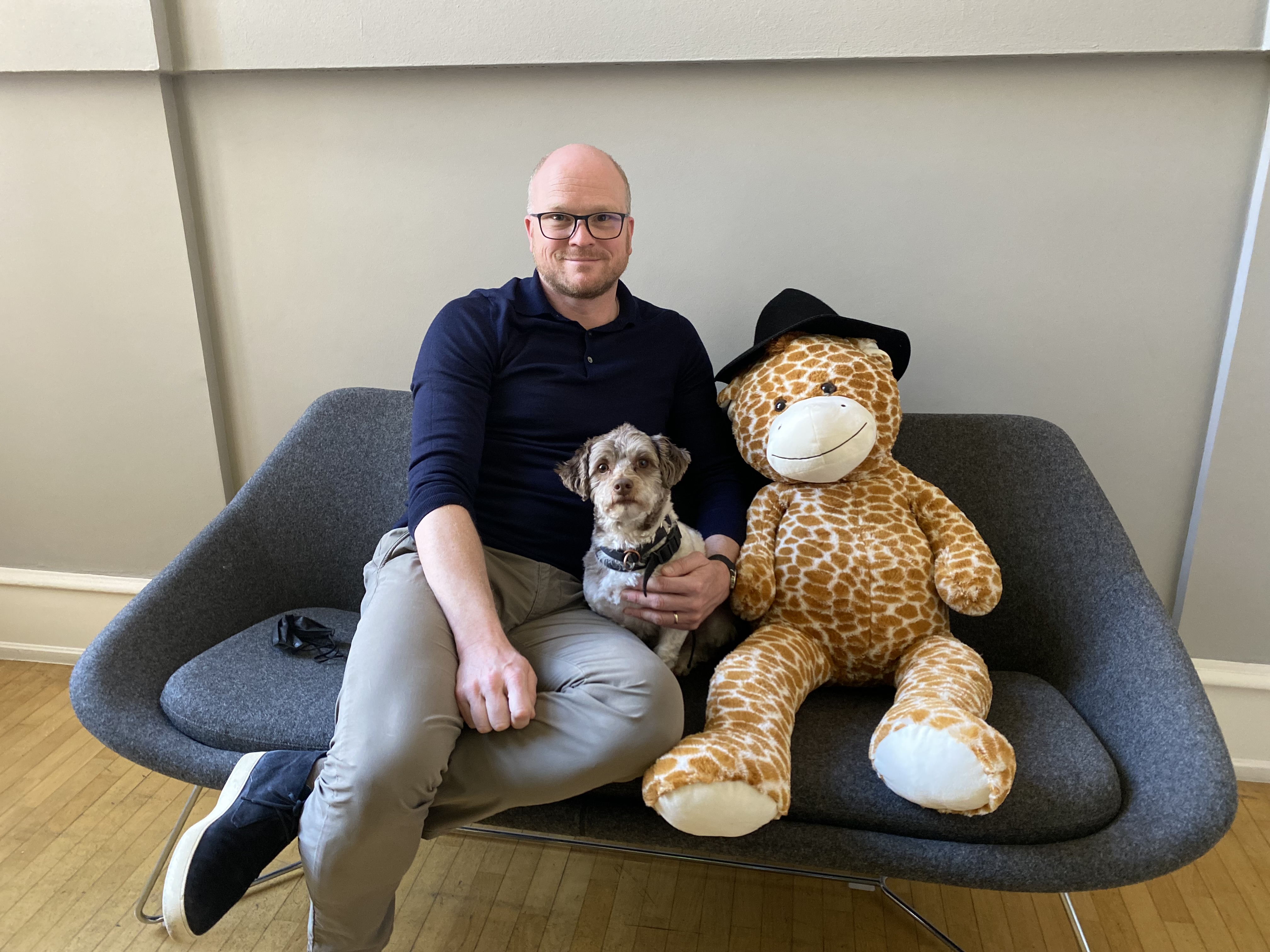 Ein Mann sitzt mit einem kleinen Hund und einer Stofftiergiraffe auf einem grauen Sofa.