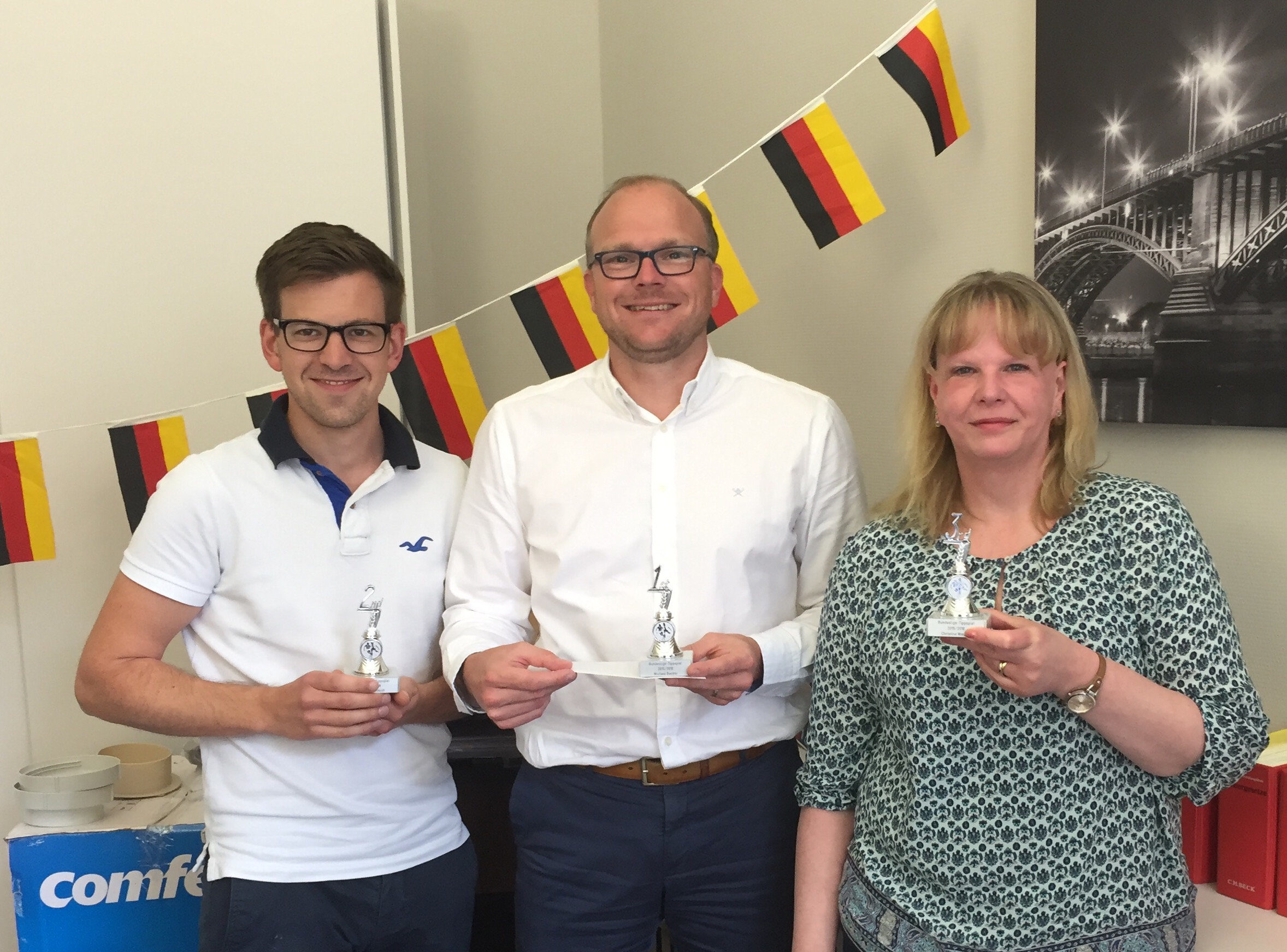 Zwei Männer und eine Frau stehen vor einer Wand, an der eine Deutschlandflaggen-Girlande hängt und halten kleine Pokale