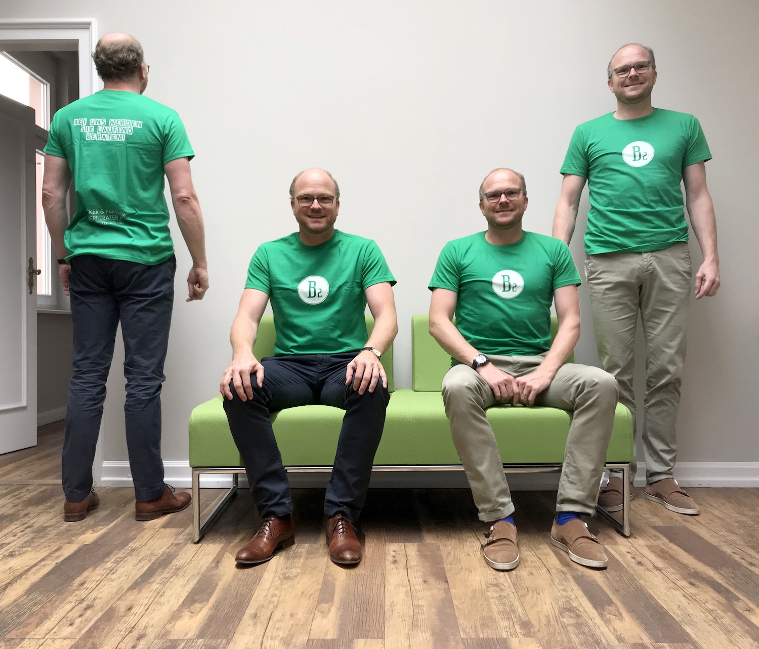 Vier Männer tragen das gleiche grüne T-Shirt. Zwei sitzen in der Mitte auf einem Sofa, zwei stehen daneben.