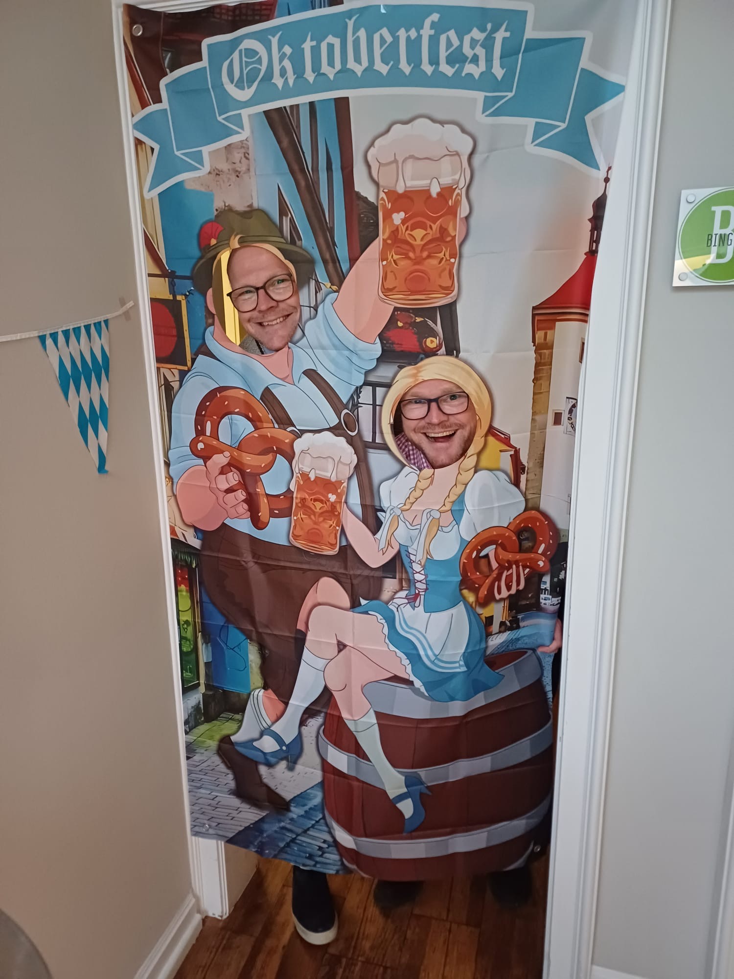 Hinter einem Oktoberfestaufsteller sind durch Ausschnitte die Gesichter der Geschäftsführer zu sehen.
