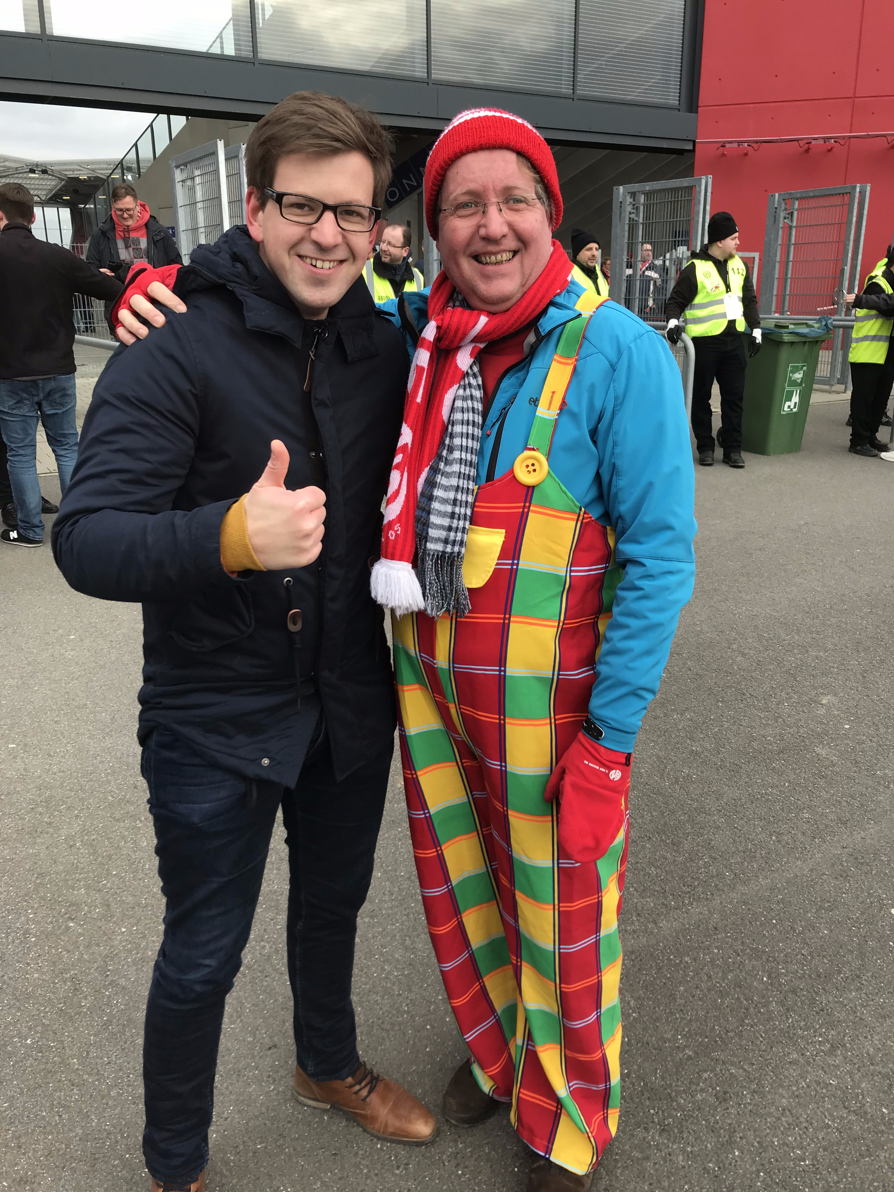 Zwei Männer stehen nebeneinander. Ein Mann trägt ein buntes Kostüm, der andere zeigt einen Daumen nach oben.
