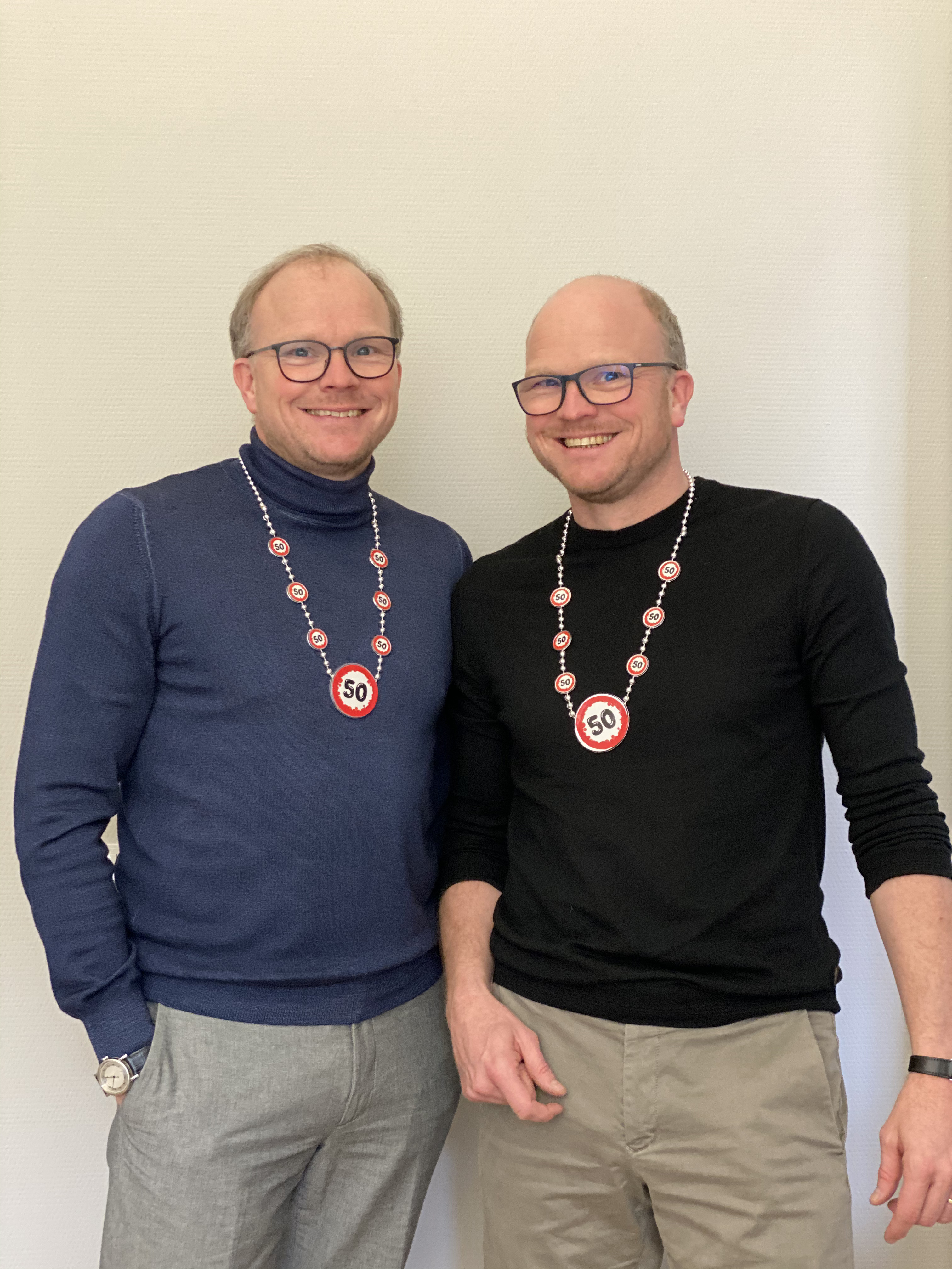 Die Geschäftsführer Michael und Stefan Becker lächeln in die Kamera und tragen Ketten mit einem Anhänger auf dem 50 steht.