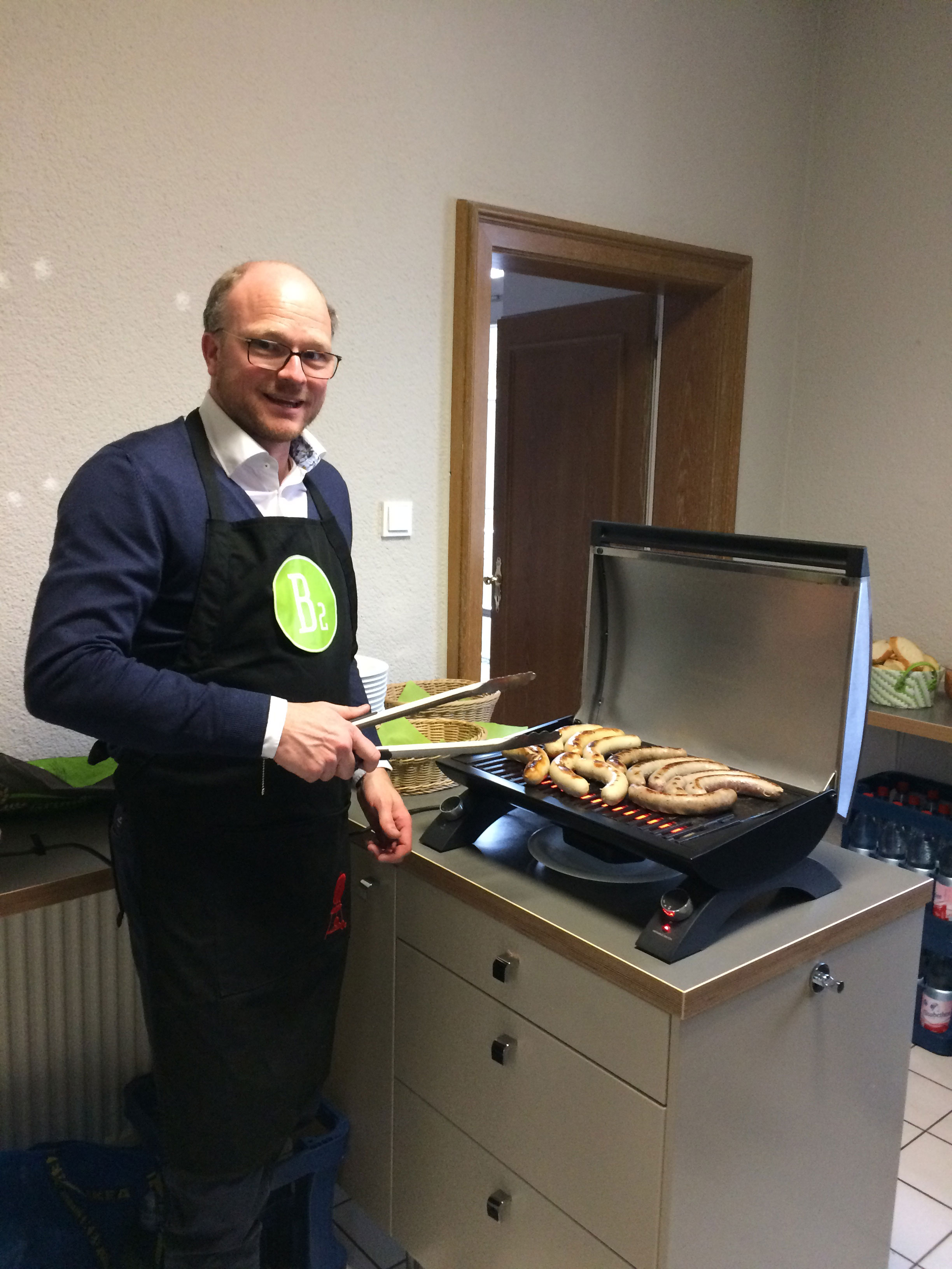 Einer der Geschäftsführer steht vor einem Tischgrill und lächelt in die Kamera.