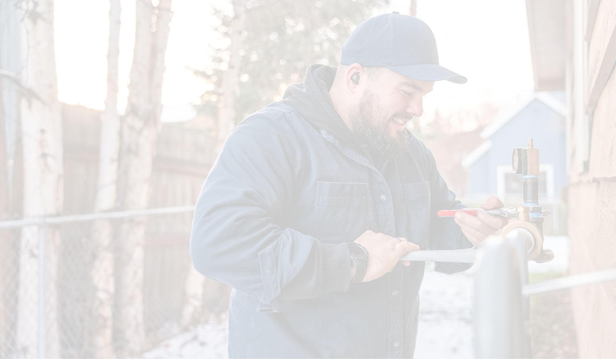Smiling man in a blue jacket and cap using a wrench to adjust outdoor plumbing pipes near a house.