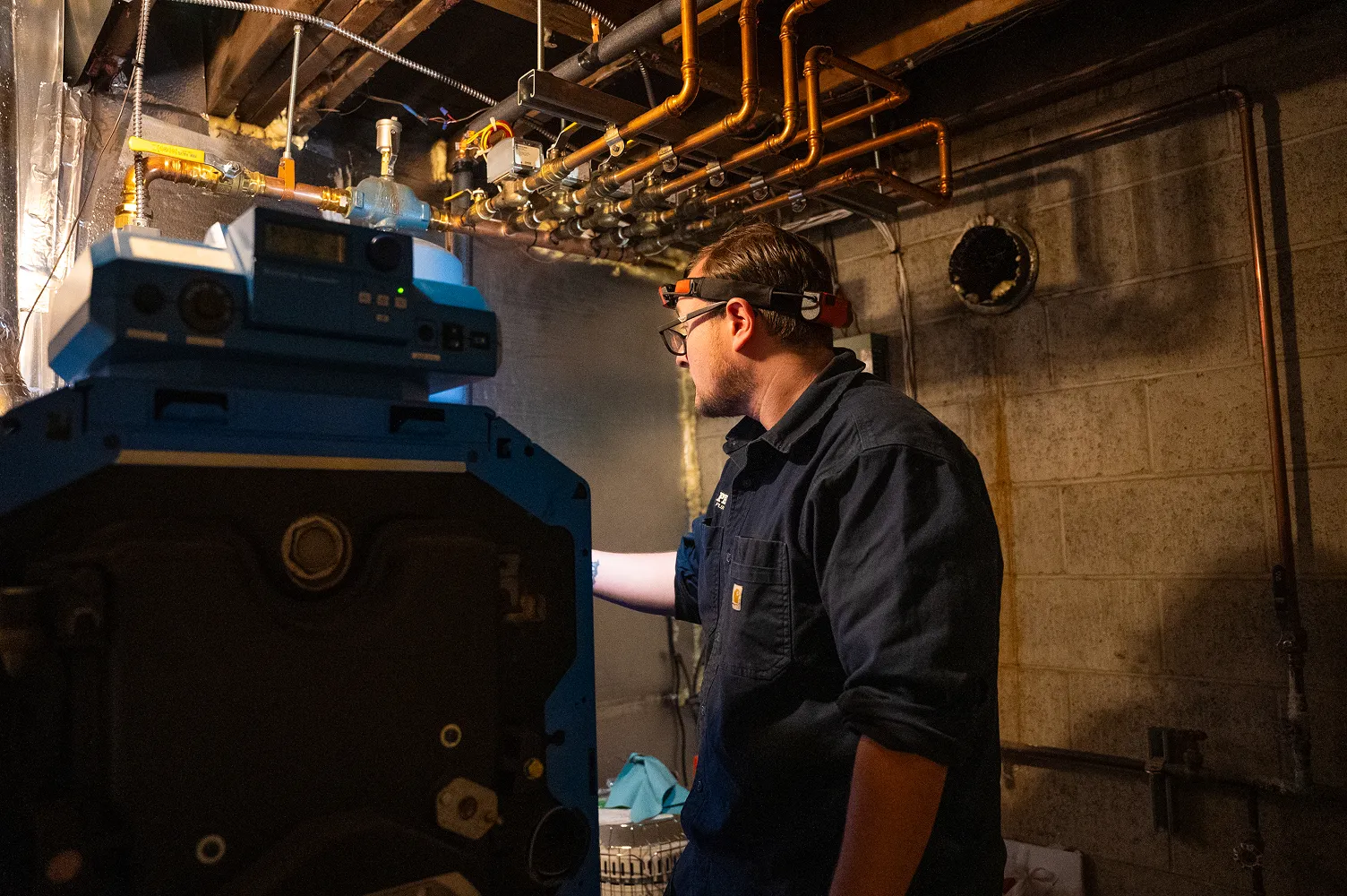 Technician wearing glasses and a headlamp inspecting plumbing pipes and equipment in a dimly lit basement.