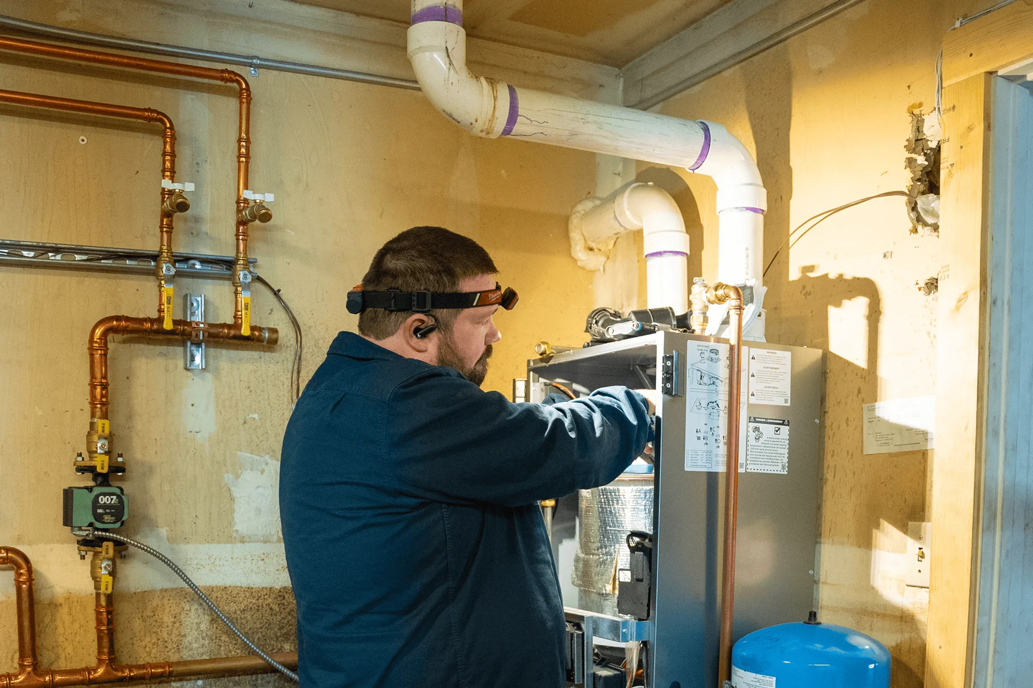 Technician in a blue jacket working on a furnace or HVAC unit inside a utility room with copper pipes and white ventilation ducts.