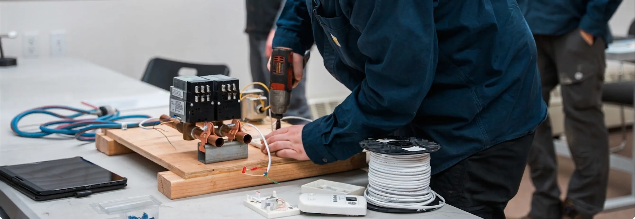 Technician using a power drill on a wooden board with electrical components and coiled wire on a table.