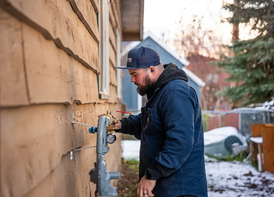 Man in a navy jacket and cap adjusting a gas meter on the side of a wooden house.