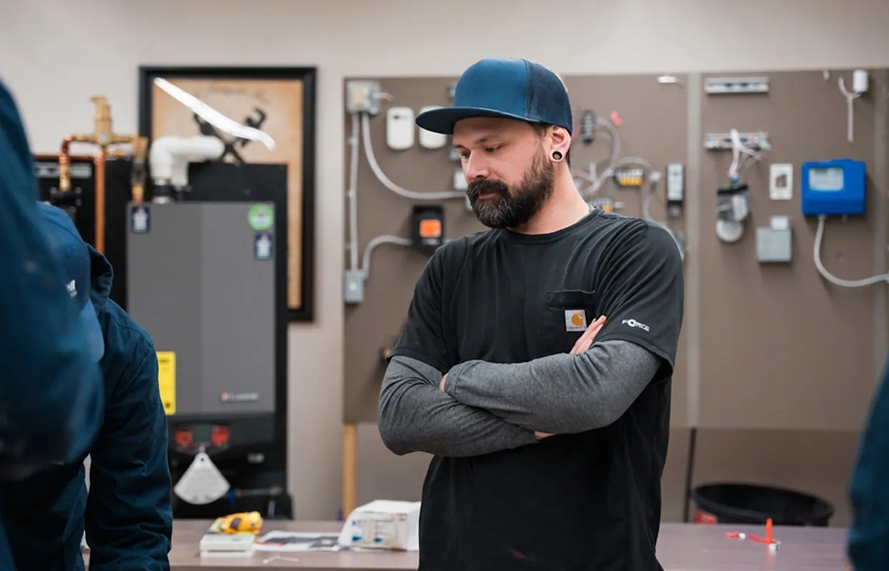 Man with a beard wearing a blue cap and black shirt with folded arms in a workshop with electrical equipment on the wall behind him.