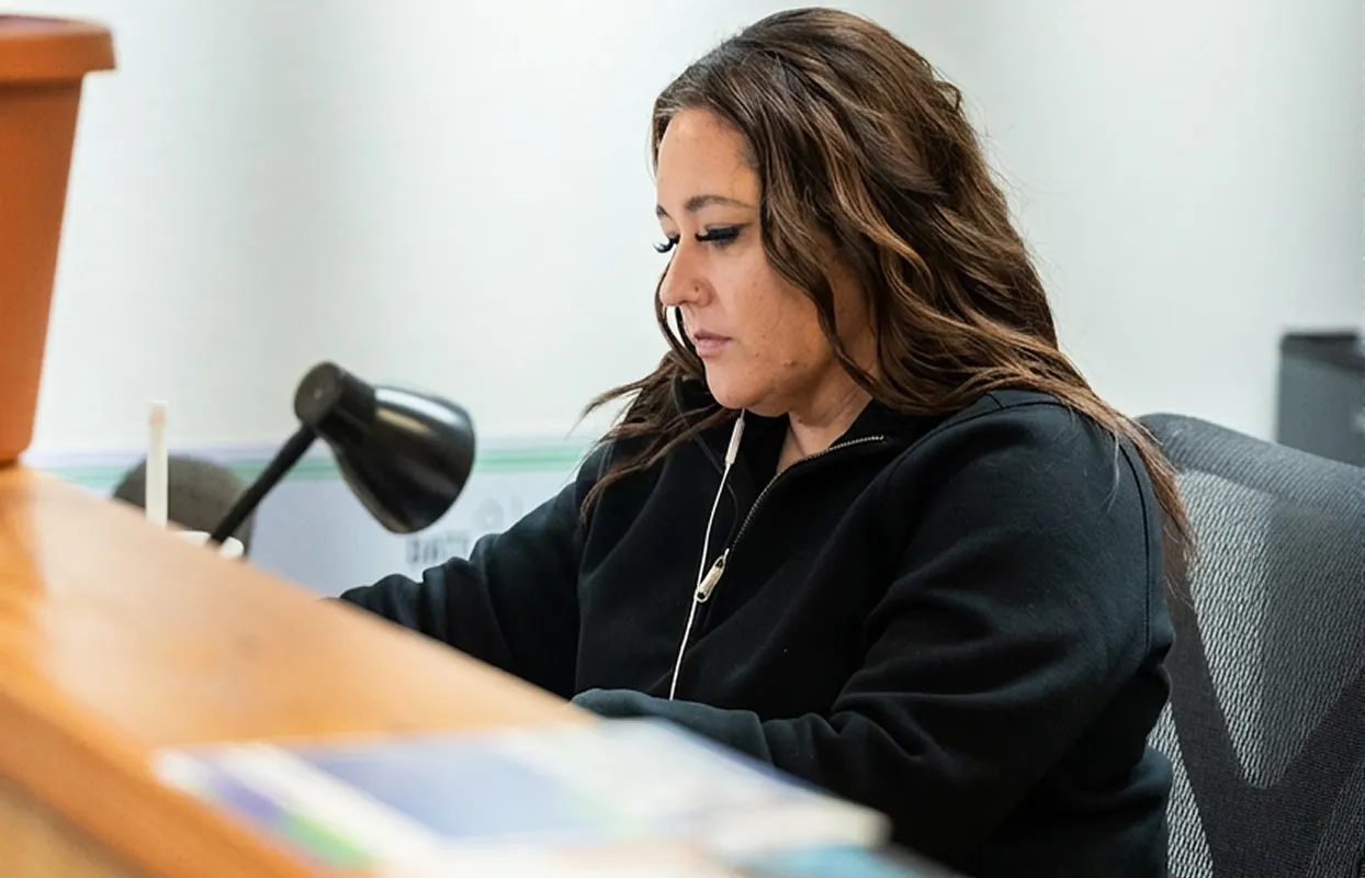 A woman with long wavy hair wearing a black zip-up jacket, sitting and concentrating while using a device with earphones.