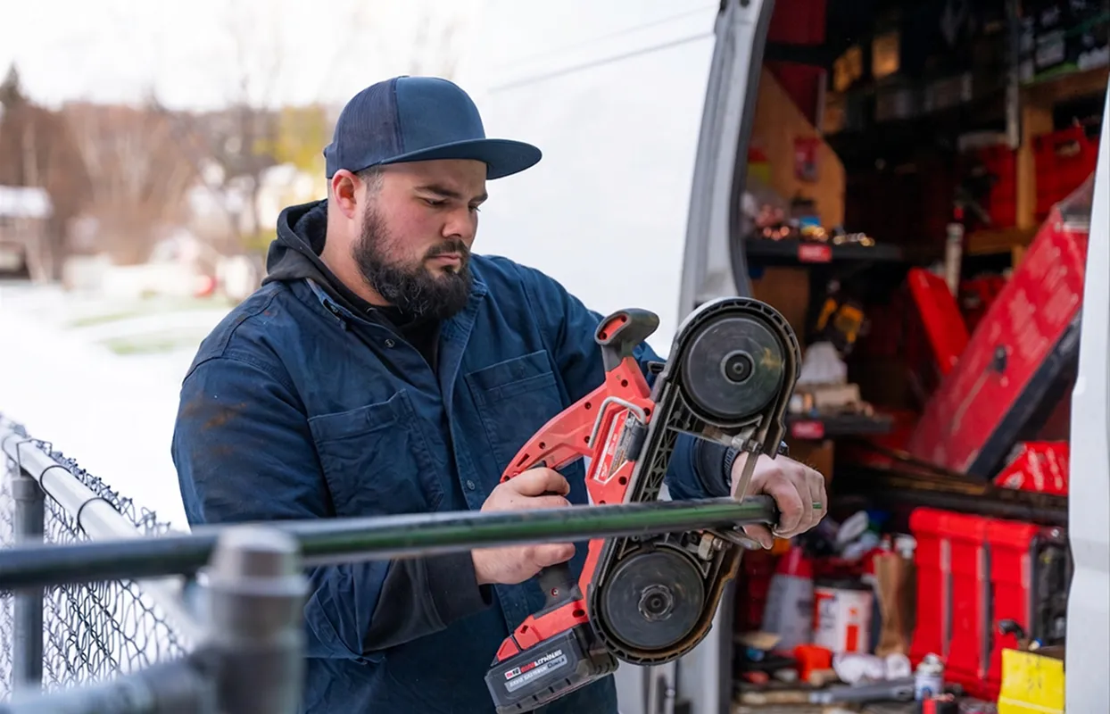 Man in blue jacket and cap using a red power tool to cut a metal pipe near a vehicle with tools inside.
