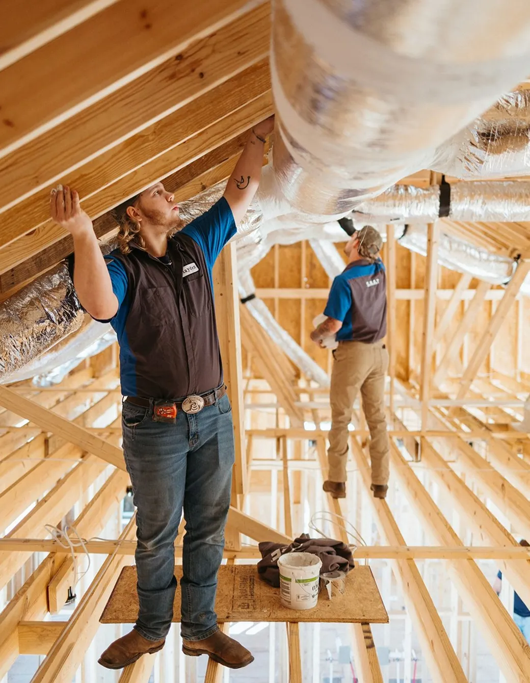 Two construction workers installing insulated ductwork in a wooden attic framework.