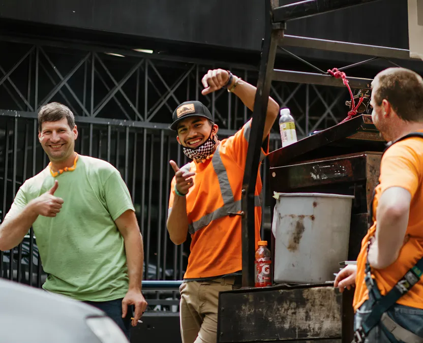 Three male workers smiling and gesturing thumbs up and pointing while standing near a truck with tools and bottles.