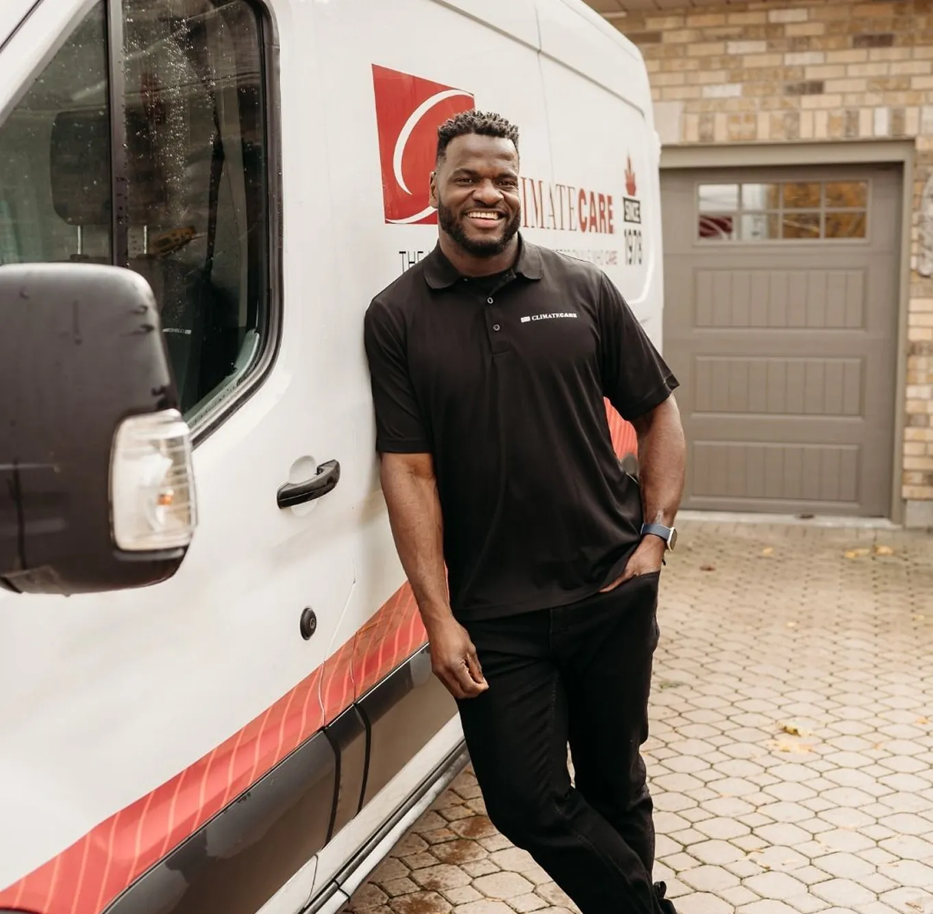 Smiling man in a black ClimateCare polo shirt leaning against a white ClimateCare service van parked on a driveway.