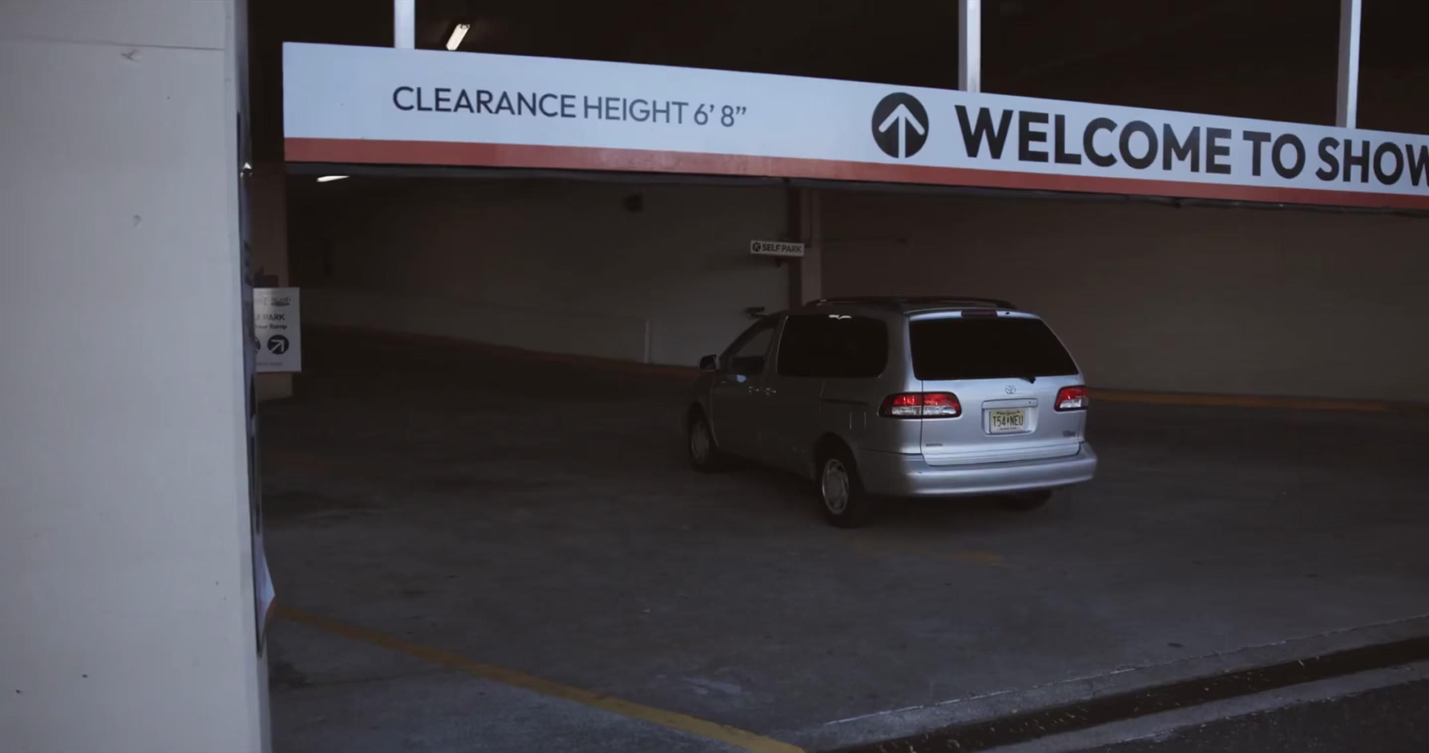 Entrance to a self-park parking garage with a clearance height sign reading 6 feet 8 inches and a welcome banner, with a silver minivan driving through the entryway.