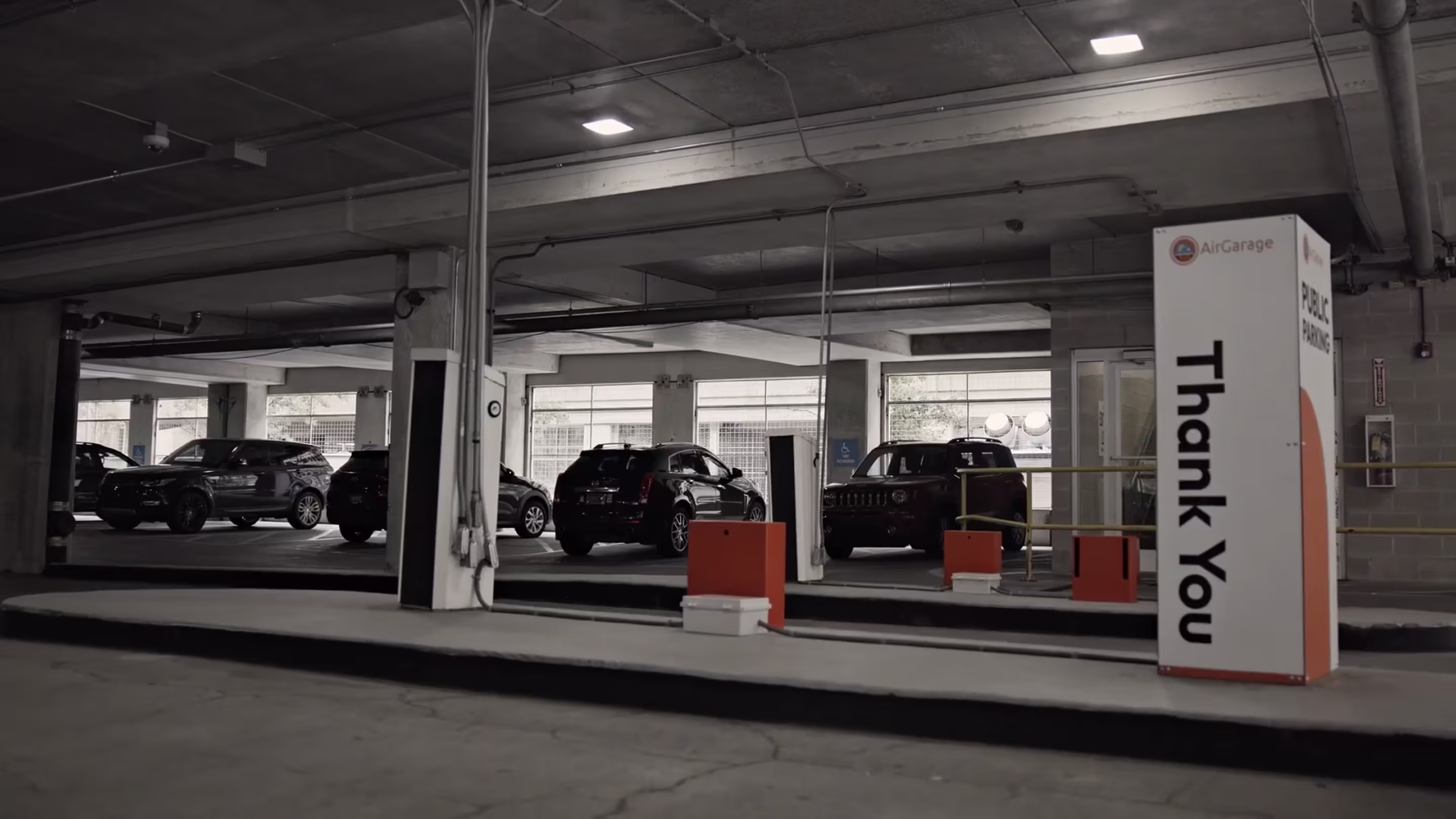Interior of a public parking garage exit with several parked SUVs, orange gate arms, and an AirGarage sign reading 'Thank You' and 'Public Parking' at the exit lane.