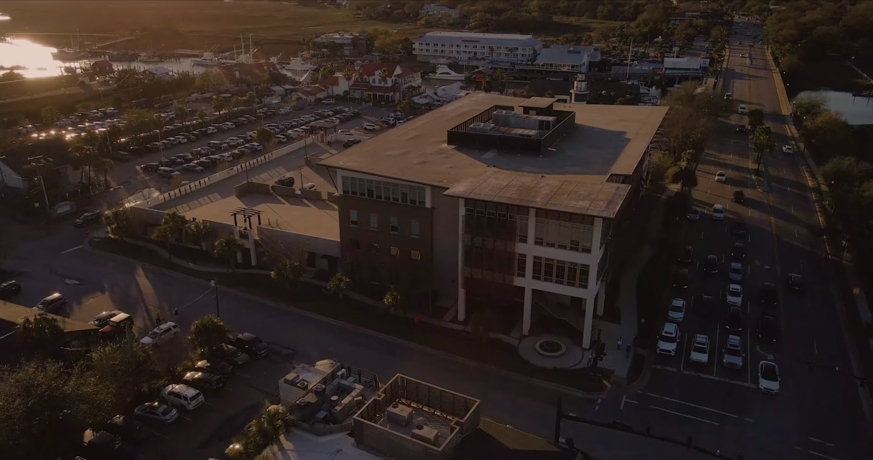 Aerial drone view at sunset of a coastal waterfront town, showing a large parking lot next to a multi-story building, a marina with docked boats, and streets lined with parked cars and palm trees.