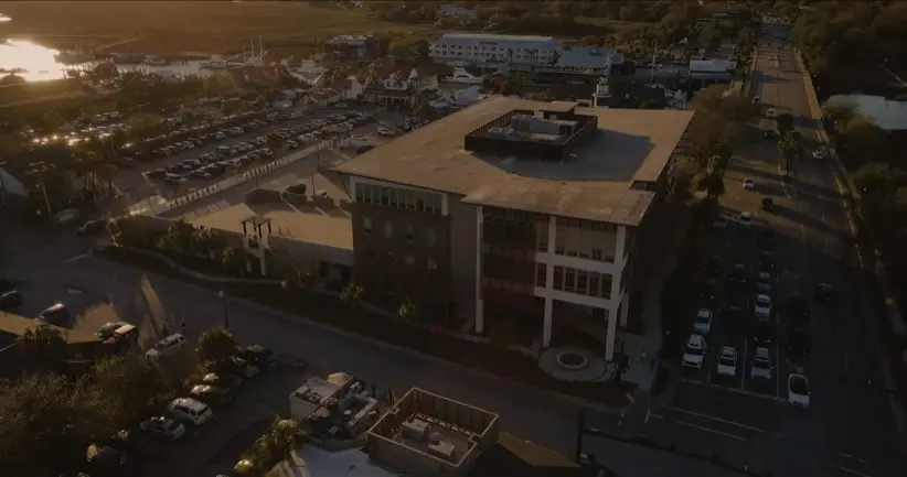 Aerial drone view at sunset of a coastal waterfront town, showing a large parking lot next to a multi-story building, a marina with docked boats, and streets lined with parked cars and palm trees.