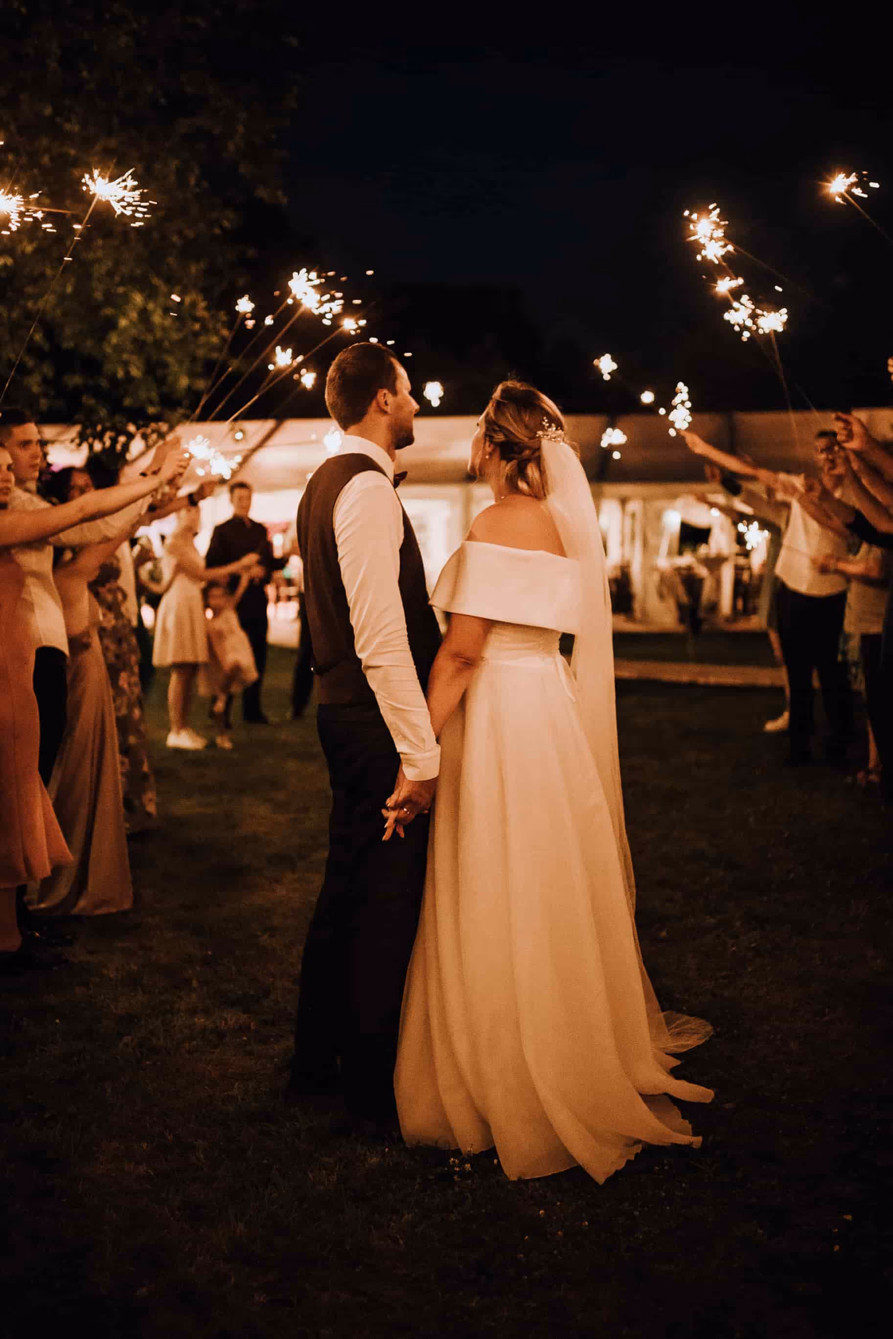Bride and groom holding hands facing each other at night, surrounded by guests holding sparklers.