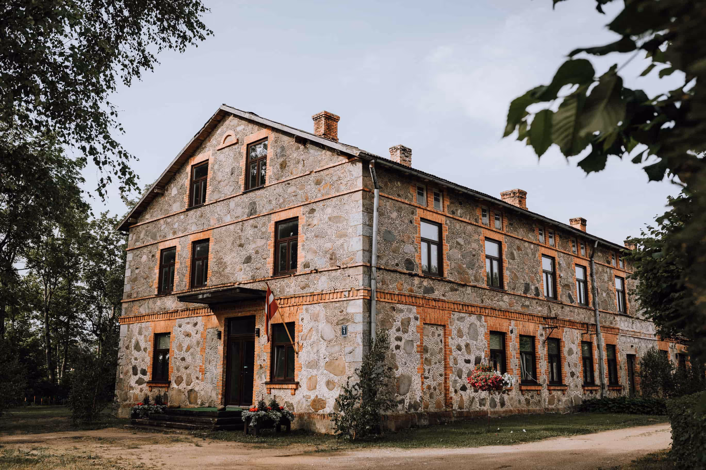 Large two-story stone building with brick window frames and a Latvian flag near the entrance, surrounded by greenery.