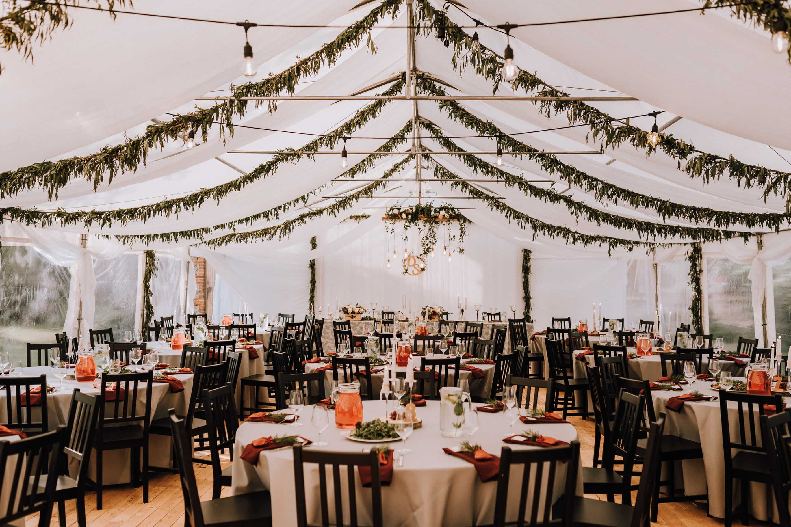 Elegantly decorated wedding reception tent with white tablecloths, black chairs, greenery garlands, and hanging lights.