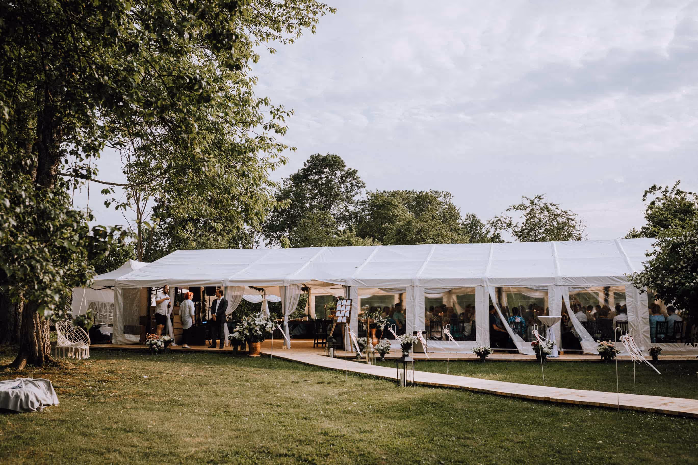 Outdoor white tent set up on grass with people inside and greenery surrounding it under a partly cloudy sky.