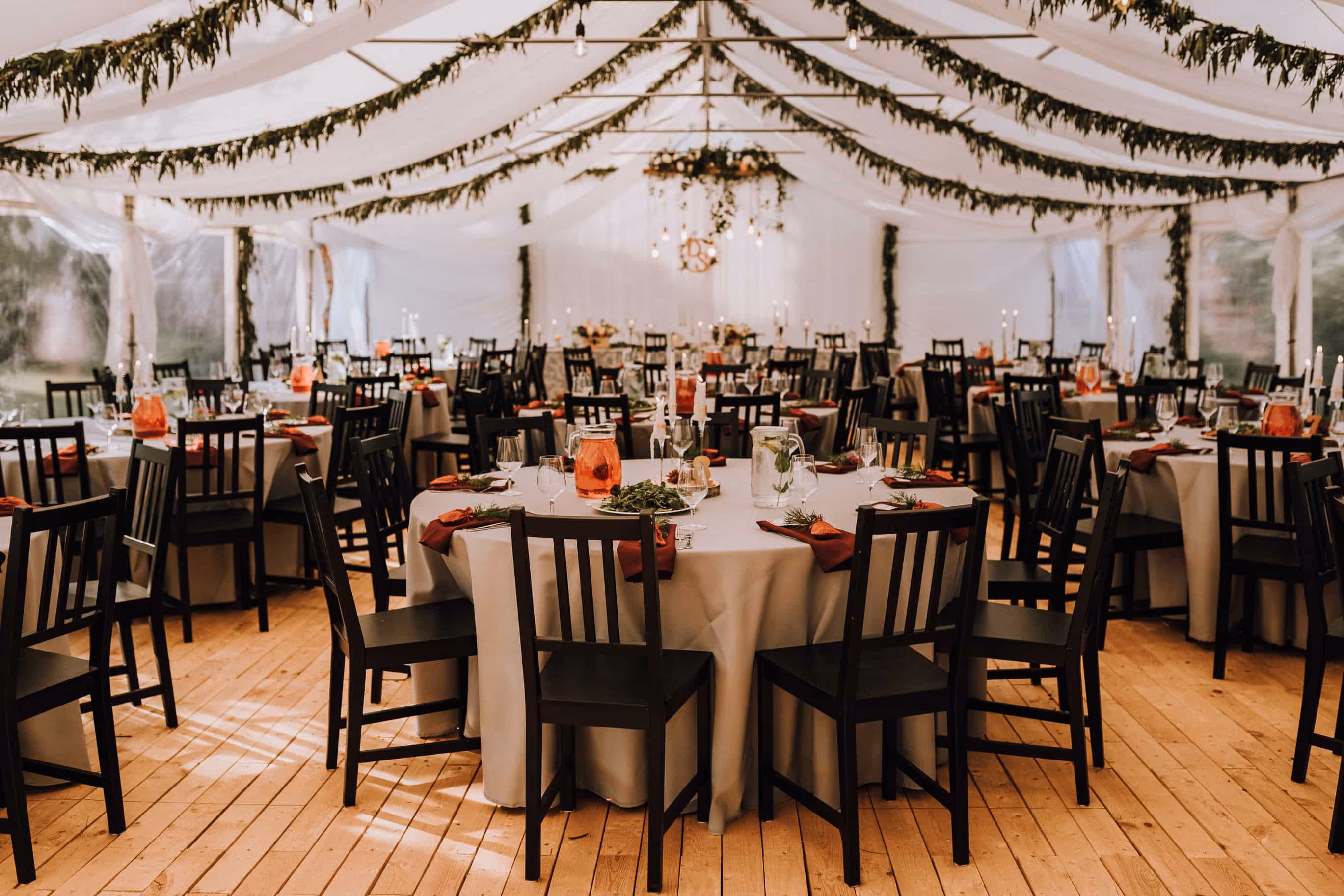 Elegant event tent decorated with garlands and round tables set with gray tablecloths, black chairs, red napkins, and pitchers of beverages.