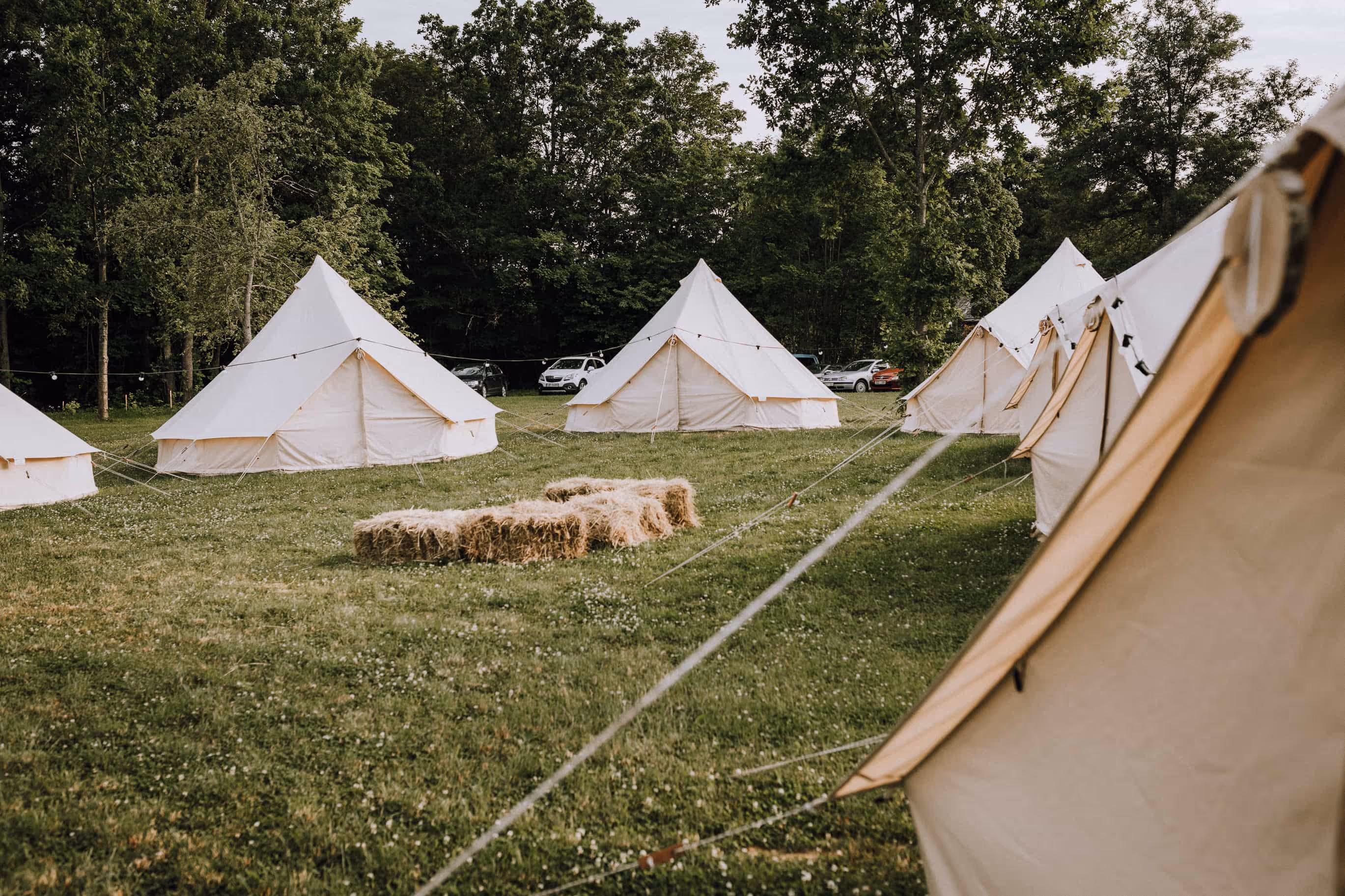 Several beige canvas tents set up on grassy field with hay bales in the center and trees in the background.