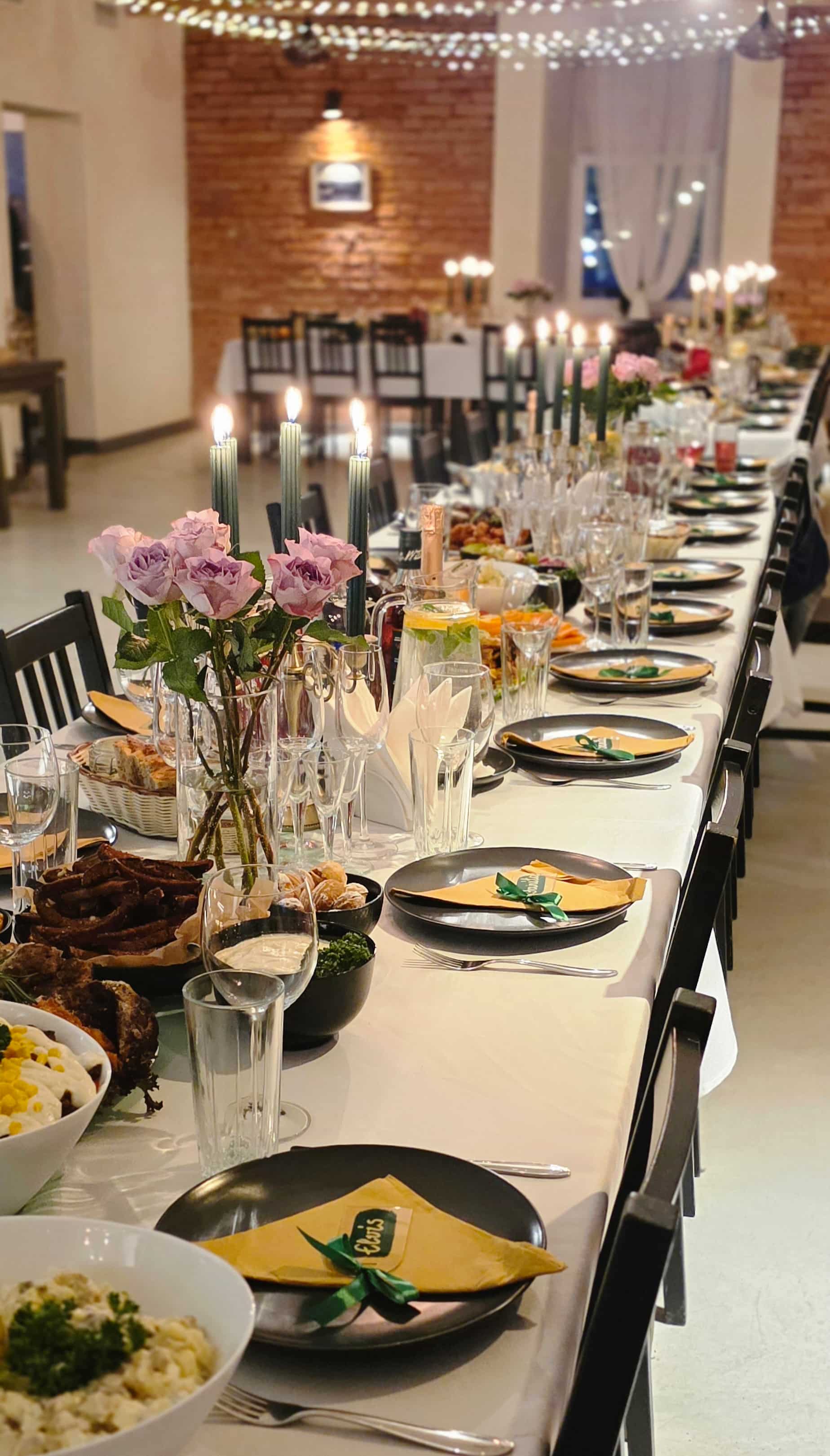 Long dining table set with black plates, yellow napkins tied with green ribbon, glassware, lit candles, pink roses, and bowls of food in a warmly lit room.