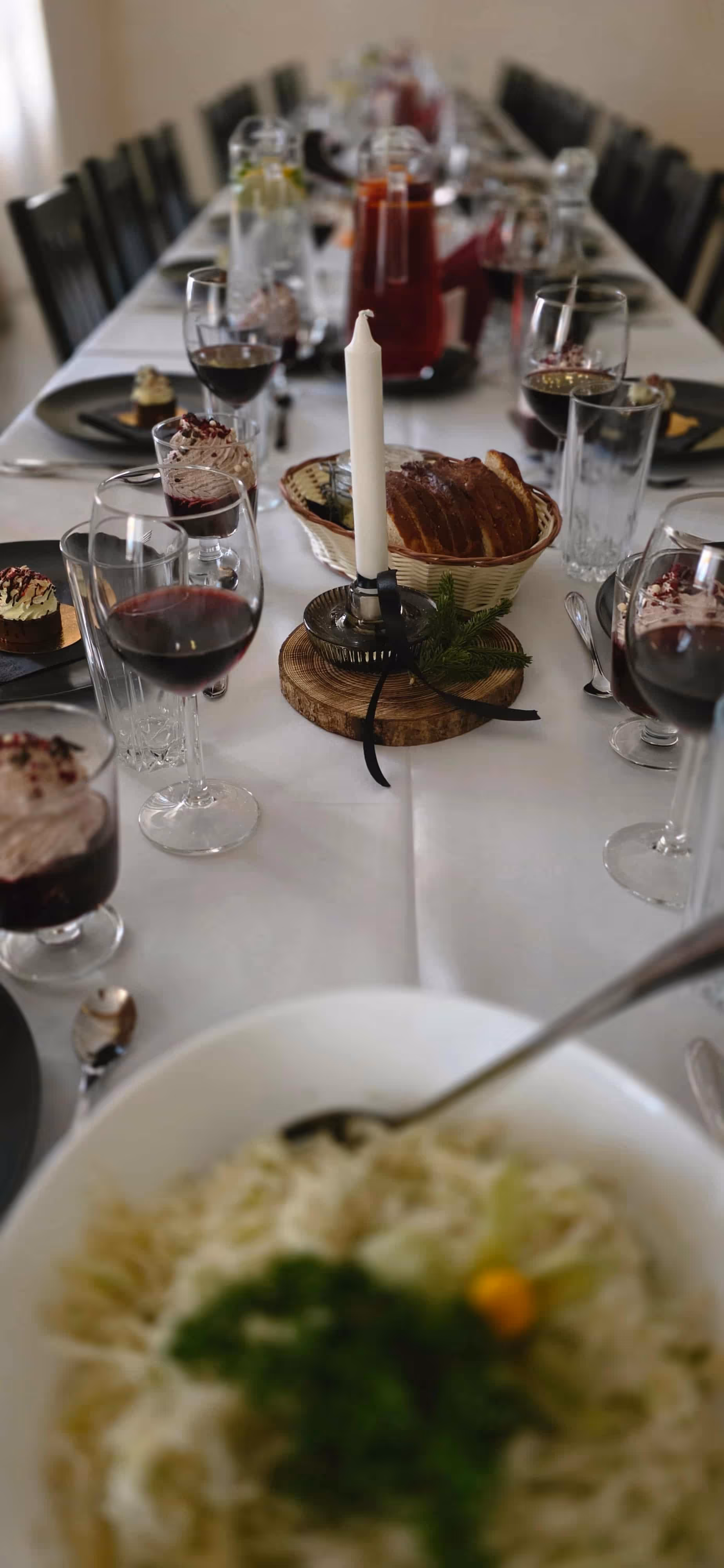 Table set for a meal with wine glasses, desserts, bread basket, a white candle centerpiece, and a bowl of pasta in the foreground.