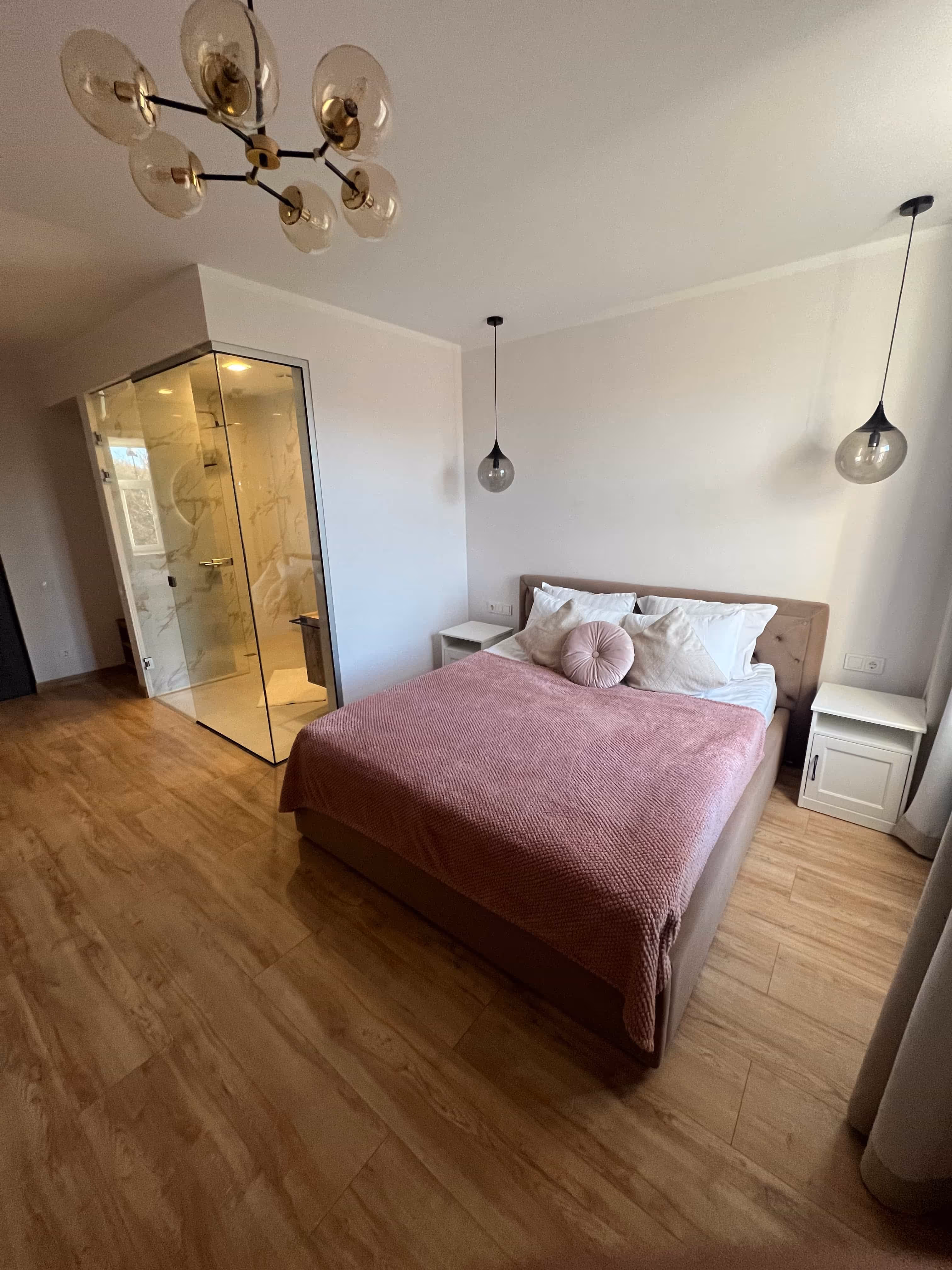 Modern bedroom with a double bed covered by a pink blanket, two hanging pendant lights, wooden floor, and a glass-enclosed shower in the corner.