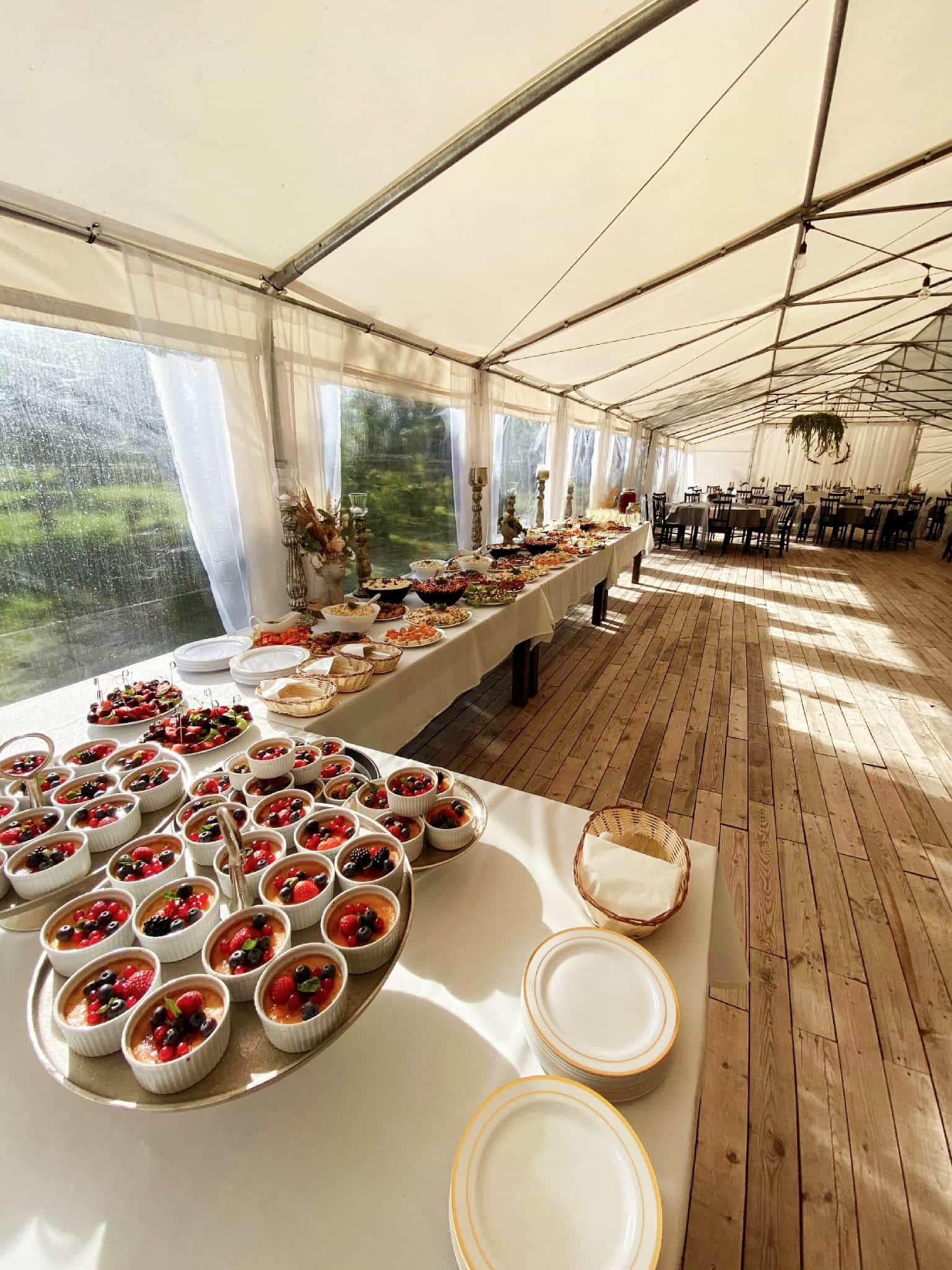 Long buffet table under a white tent with assorted desserts, fruit cups, and plates, with dining tables and chairs in the background.