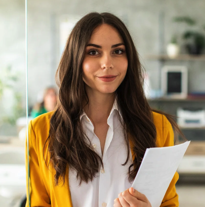 Smiling woman with long brown hair wearing a yellow blazer and white shirt holding a document in an office setting. Client picture. 