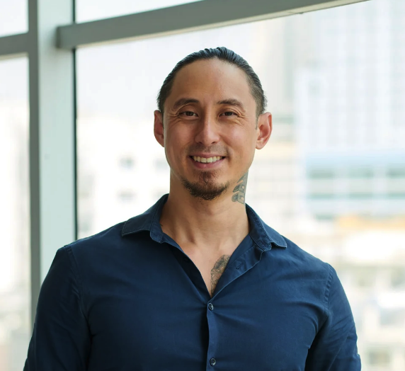 Smiling man with long hair tied back, wearing a dark blue button-up shirt, standing indoors with a blurred cityscape background. Client picture. 