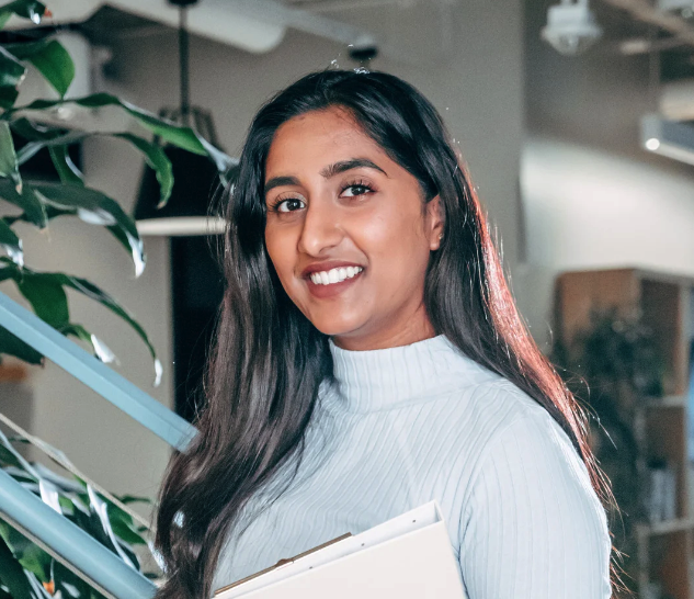 Smiling woman with long dark hair wearing a white ribbed turtleneck sweater holding a folder indoors near a plant and staircase. Client picture. 