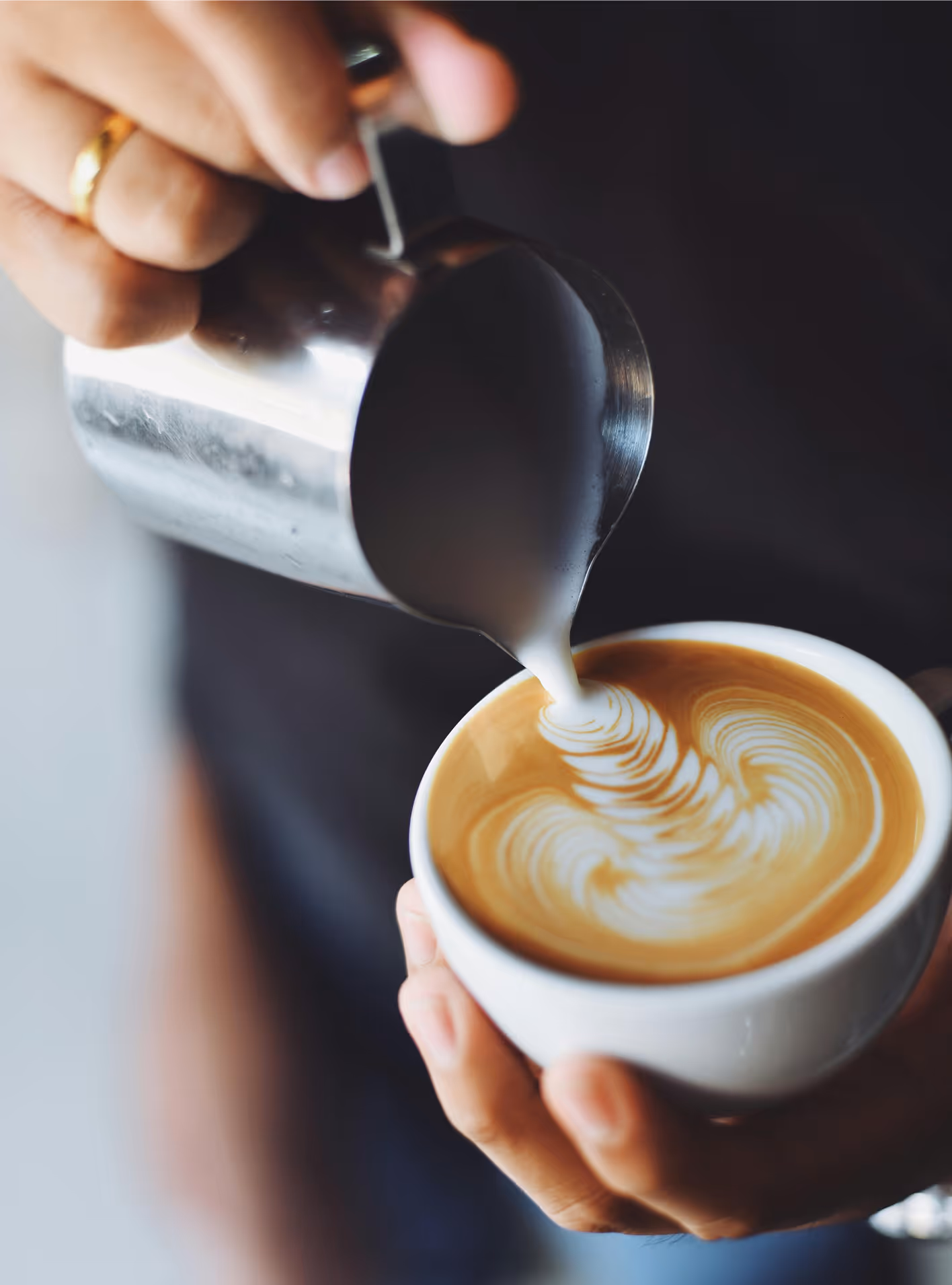 A barista pours latte into a white cup. Only the hands and latte are in view, the background is heavily blurred.