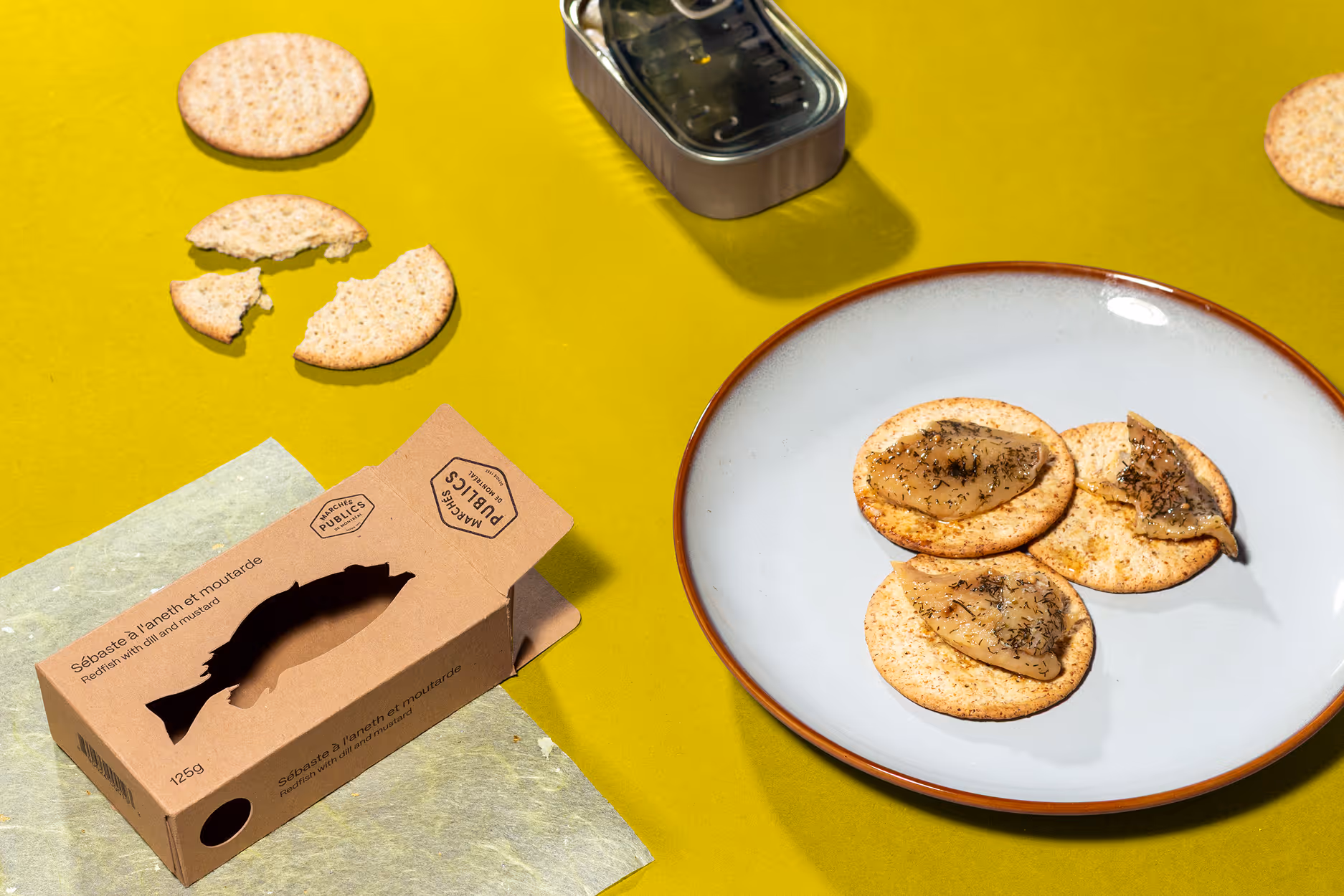 An assortment of crackers is placed on a plate, with an open packaged of tinned fish. The items are placed on a yellow background. Photo taken by © Kayleigh Lord.