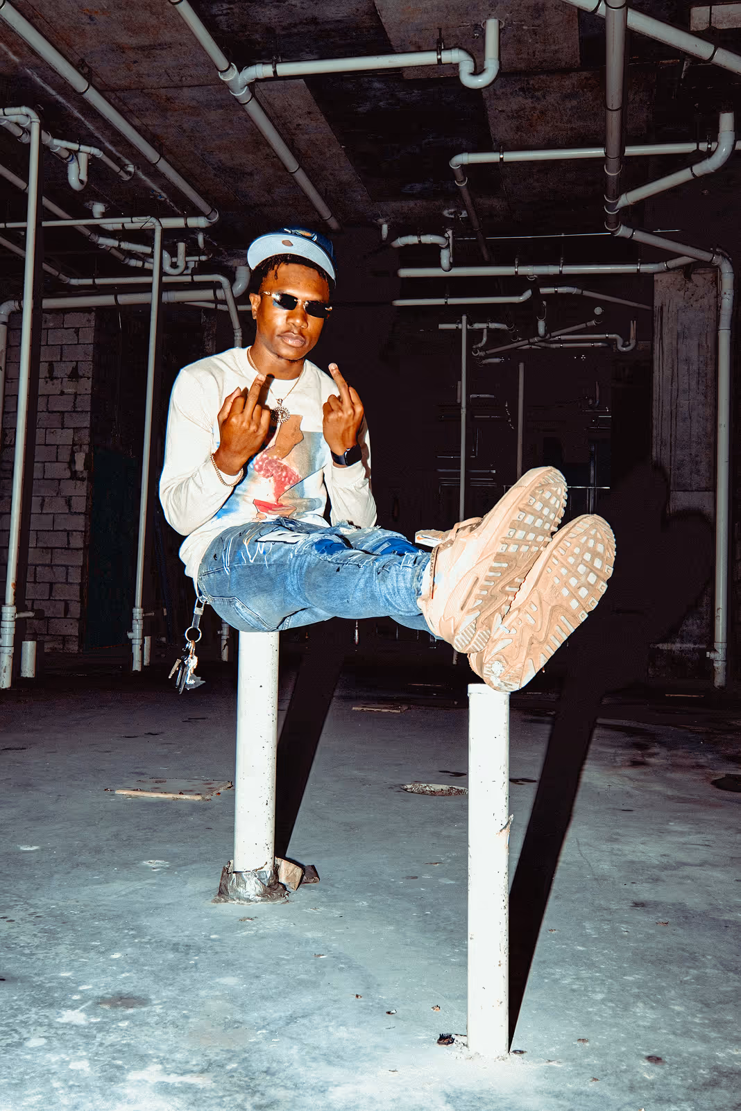 A high flash photograph of a man in casual clothes, sitting on two poles with both hands displaying the middle finger in an abandoned warehouse.