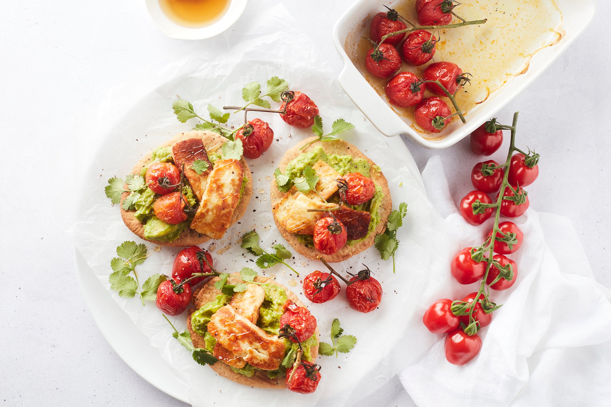 Een heldere foodfoto van mini naanbroodjes belegd met avocadocrème, gegrilde halloumi en gepofte Frieda tapastomaten, geserveerd met verse koriander en een ovenschaal met extra geroosterde tomaatjes.p