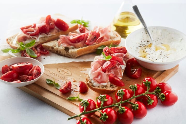 Een culinaire foodfoto van een recept: ciabatta belegd met parmaham en partjes Frieda tapastomaten, gepresenteerd op een houten serveerplank van Hoogstraten naast een kom romige burratadip en verse basilicum.